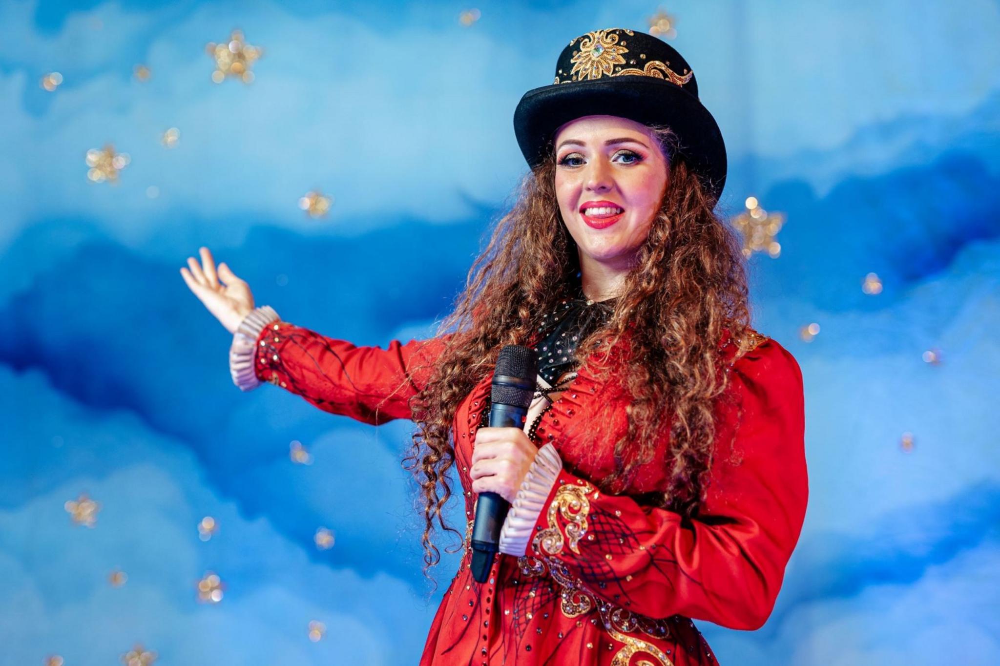 A woman with long curly brown hair performs on a stage. She wears a red coat and a black hat, while holding a microphone. She stands in front of a light blue cloud backdrop with gold stars on it.