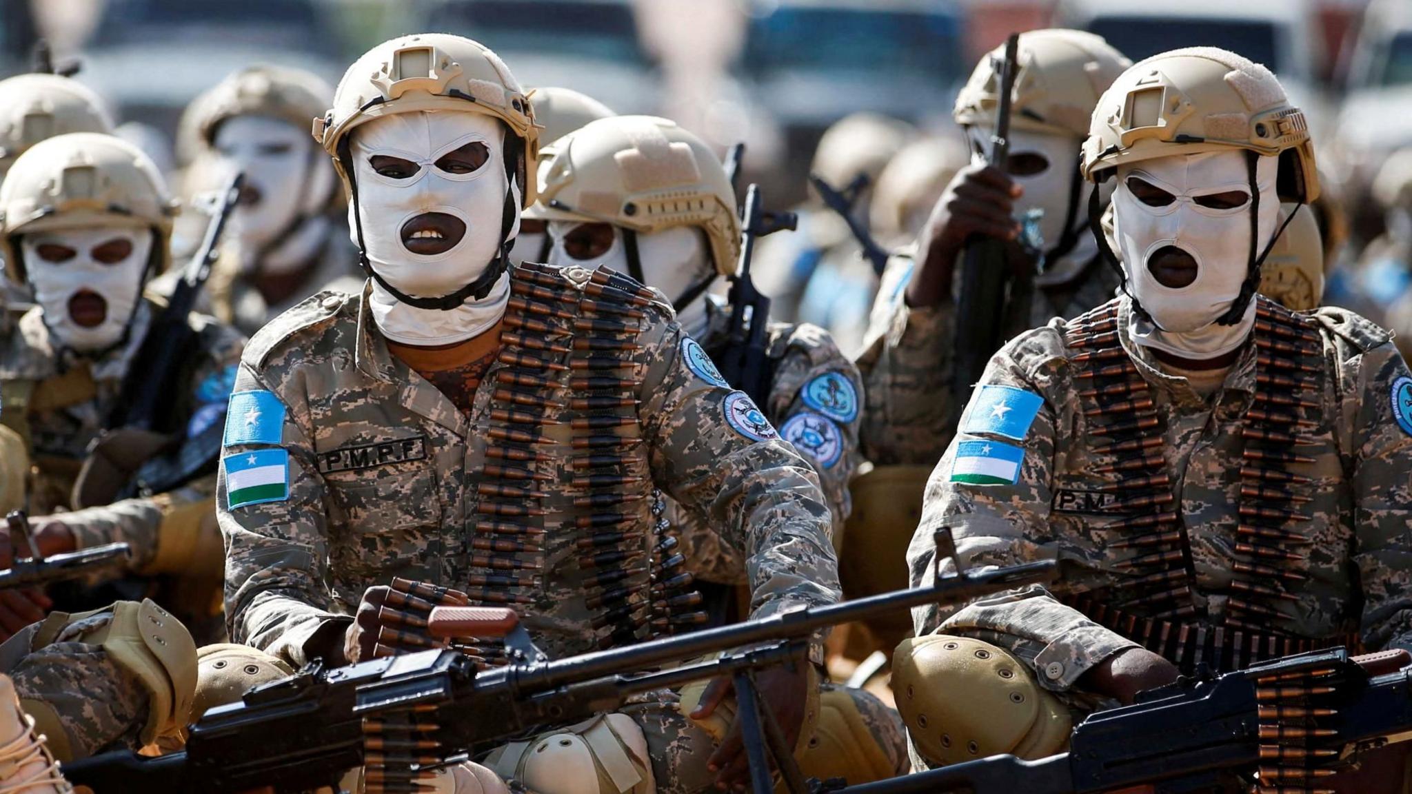 Elite Puntland soldiers sit on the ground. They are wearing white masks, beige helmets and rounds of bullets around their necks for guns they hold  