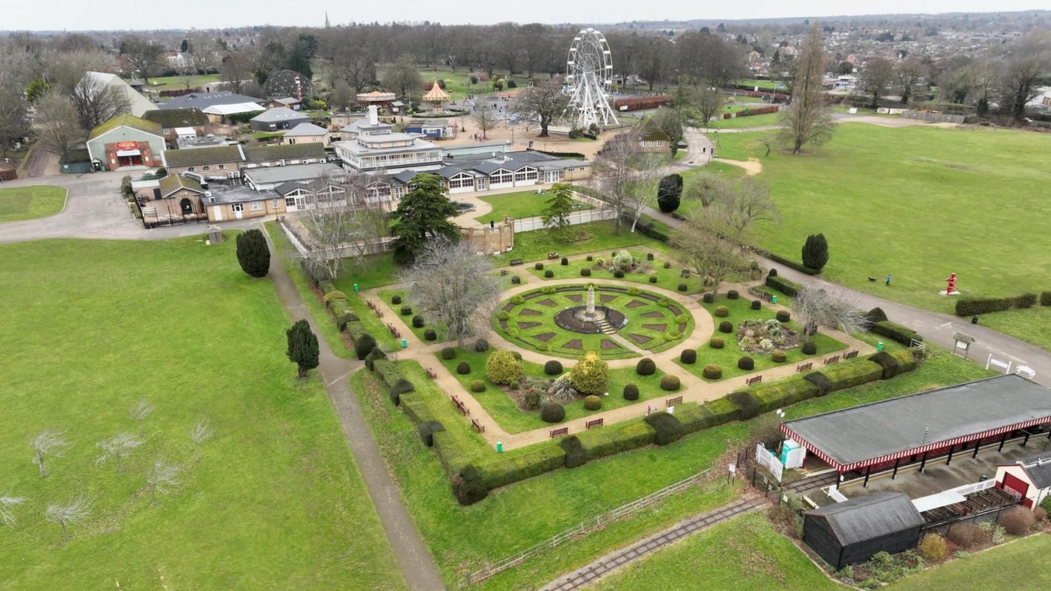 An aerial view of Wicksteed Park with ornamental garden and miniature railway track and station in the foreground. A big wheel and other buildings and rides are in the background, with grassy areas either side.