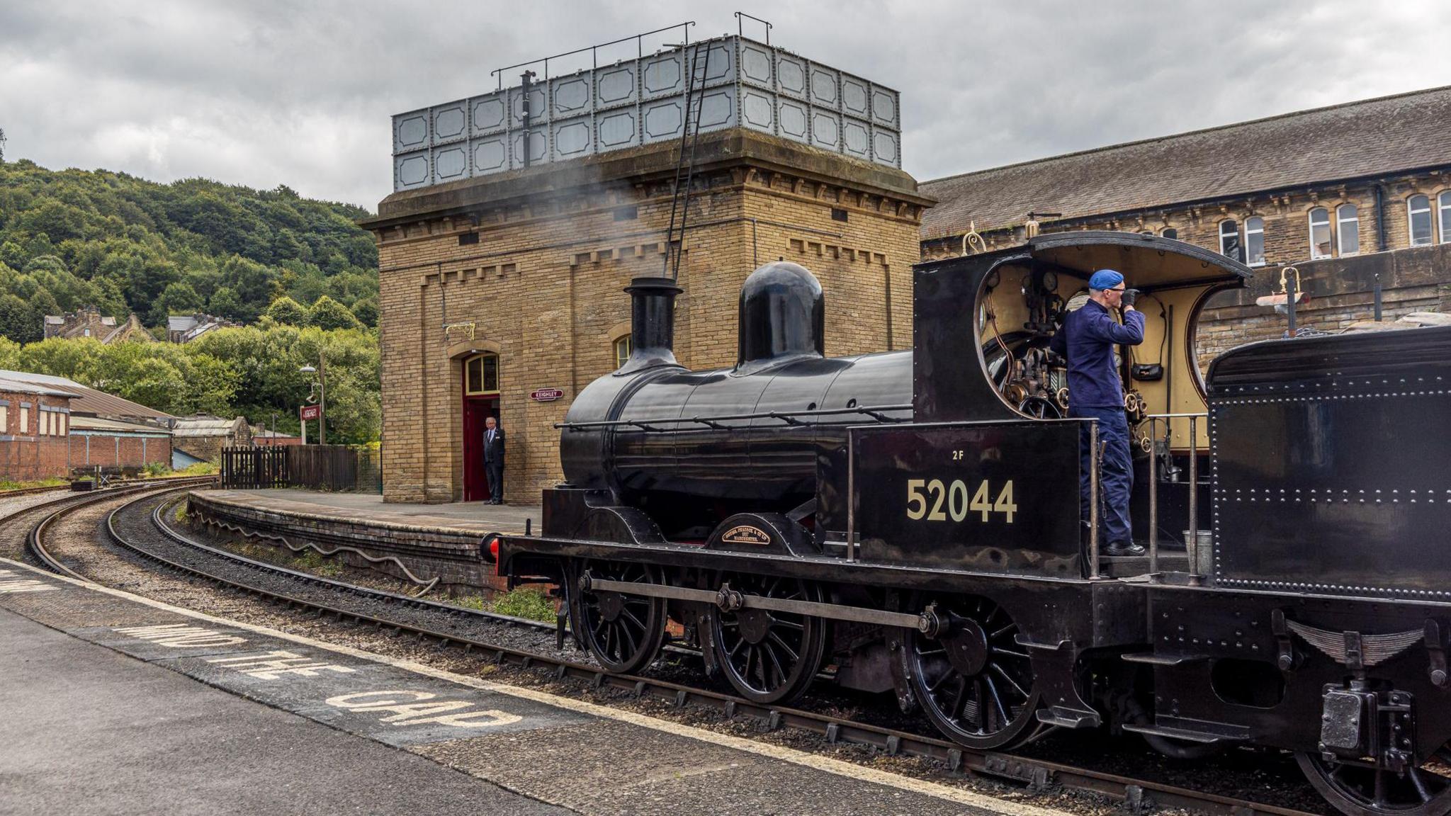 A vintage steam locomotive stationed at a railway platform. The locomotive is black and prominently displays the number 52044 on its side. A person dressed in a blue uniform and cap is standing on the footplate of the engine.
In the background, there is a brick building with a distinctive glass structure on its roof, possibly a water tower or signal box. Behind this building, there are more stone-built structures. The railway tracks curve away to the left, and the platform edge is visible in the foreground with the words "MIND THE GAP" painted in white.