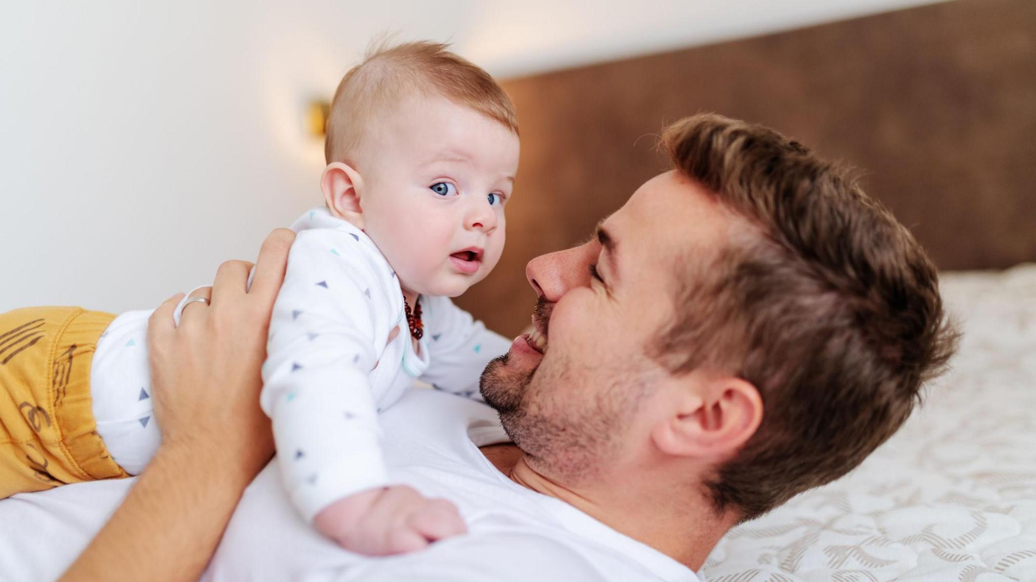 A man is lying on a bed holding a baby. The baby is looking at the camera, while the dad is looking at the baby.