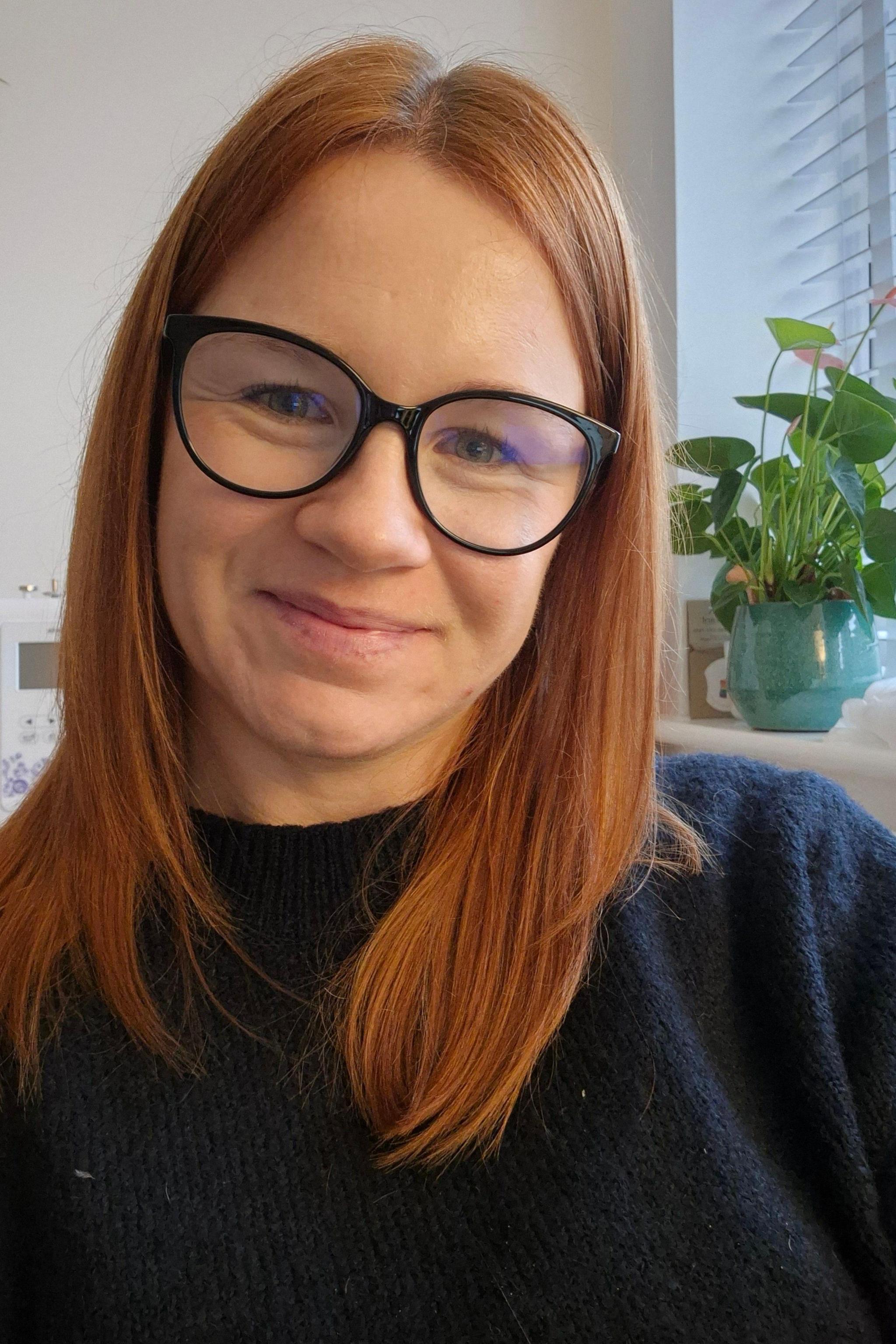 Hayley Rawlings smiles at the camera. She has shoulder-length red hair and wears a blue knitted jumper. She has black-framed glasses on. It is a selfie, and a head and shoulders shot.
