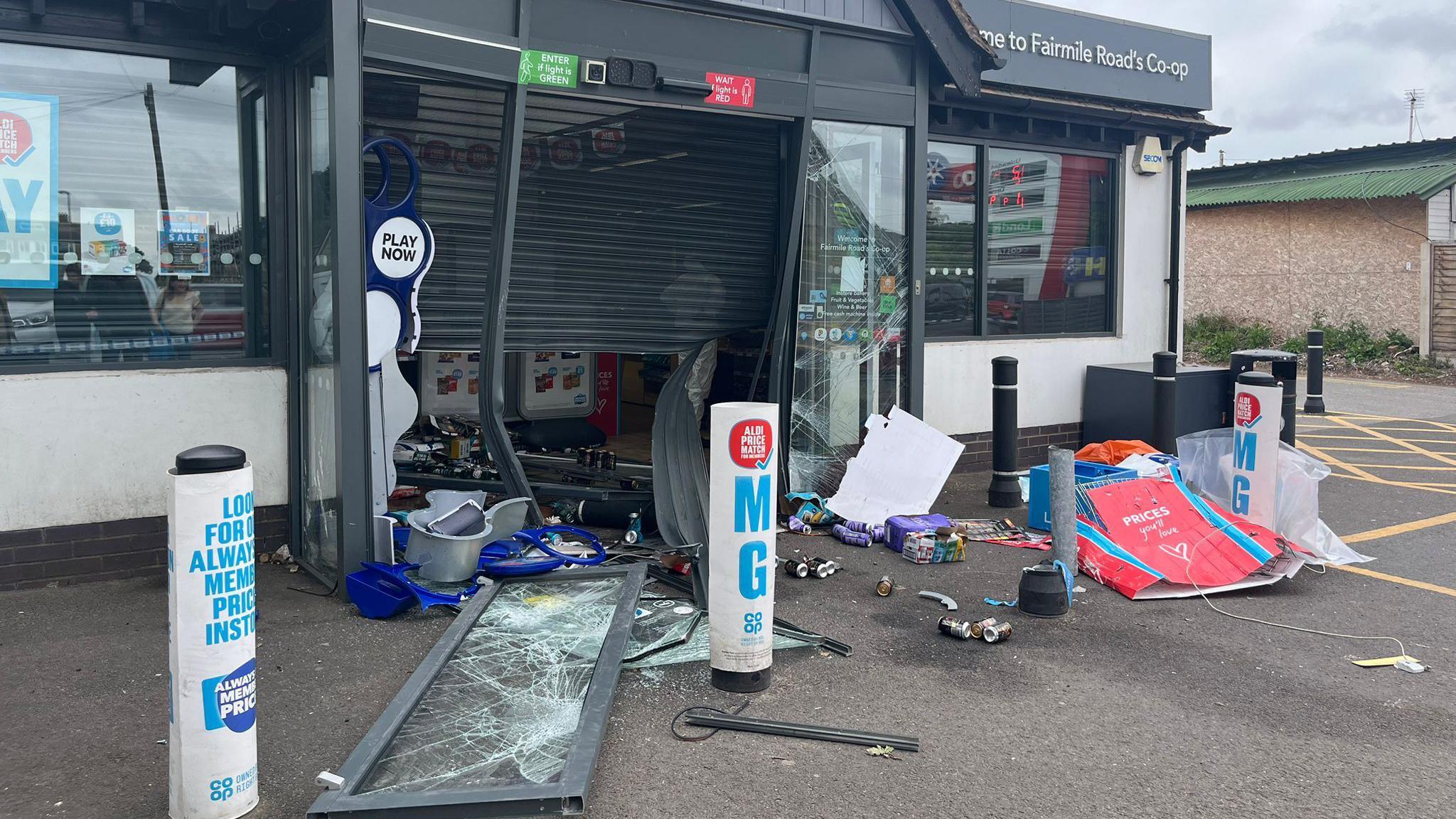 A Co-op shopfront has been smashed. There is a door with broken glass on the floor, a half opened black shutter on the front of the store, debris, litter and rubbish is seen strewn along the floor