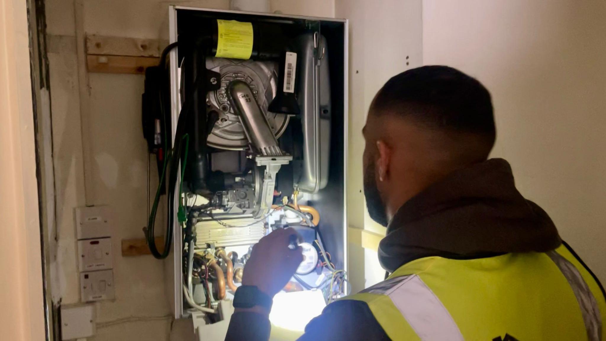 A man in a hi-vis vest shines a torch on the inner workings of a boiler.