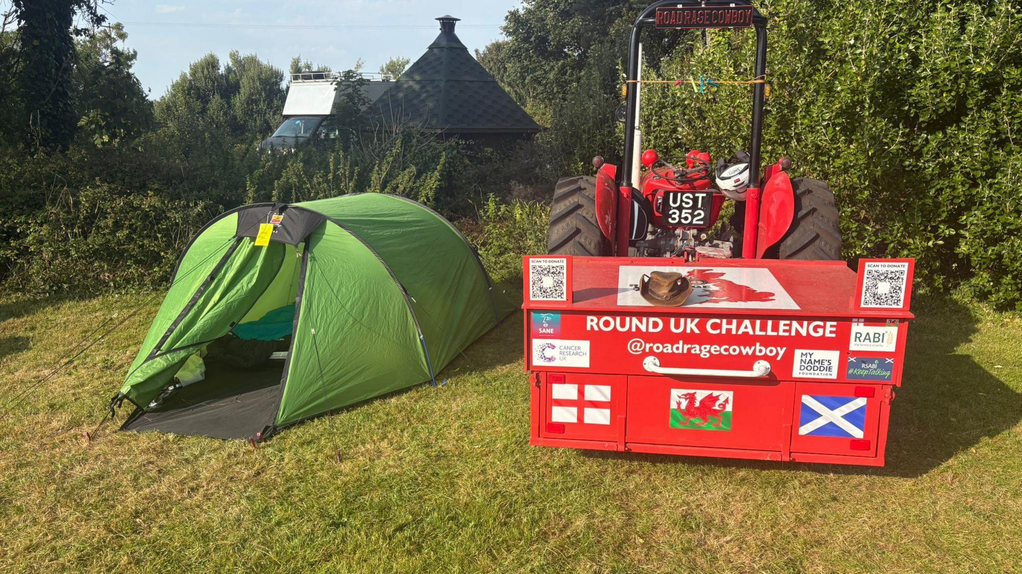 A green tent next to a the back of a red Massey Ferguson 35x tractor. The tractor has the English, Welsh and Scottish Flags and saus "Round UK Challenge @roadragecowboy". The grass is green. There are bushed and a shed in the backround. The sky is blue.