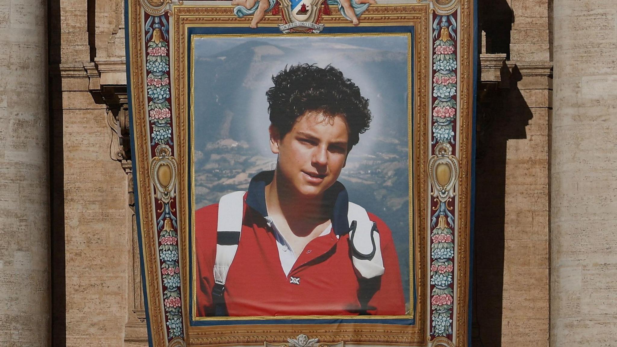 Carlo Acutis memorial, a framed photo of him with curly short black hair, wearing a red t-shirt and a white backpack. The picture is hanging from a church building outside.