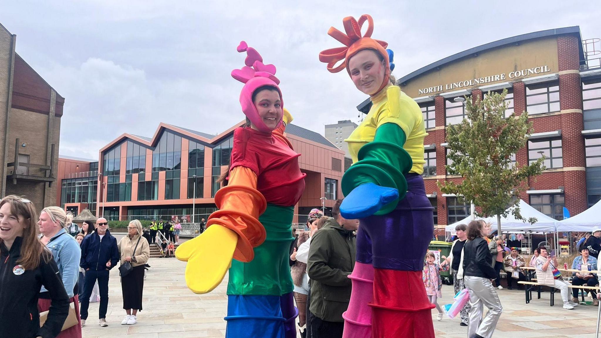 Two stilt entertainers mingle with the crowd, they are wearing rainbow coloured costumes.