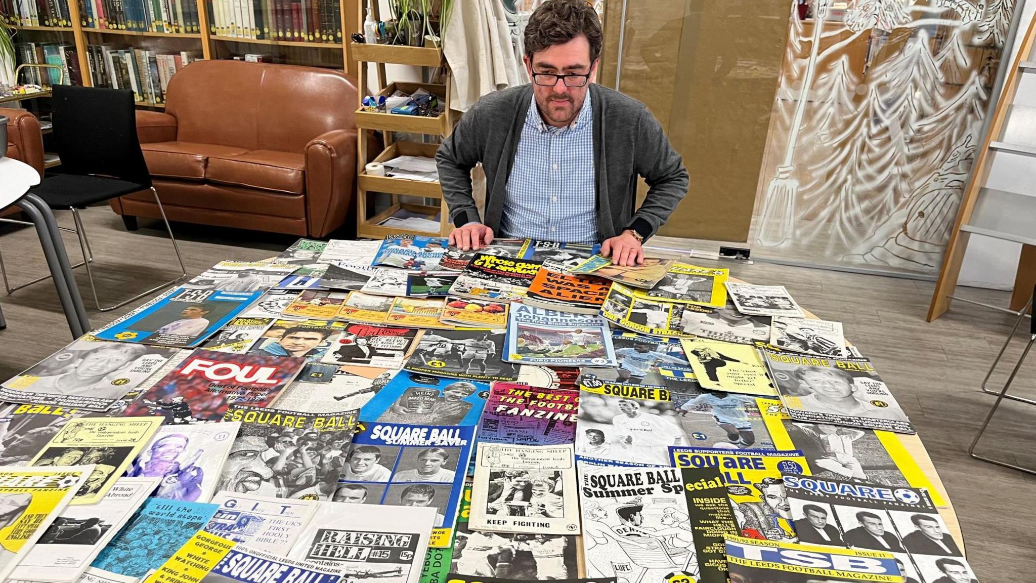 A picture of librarian Antony Ramm who is looking over dozens of football fanzines spread out on a table. 