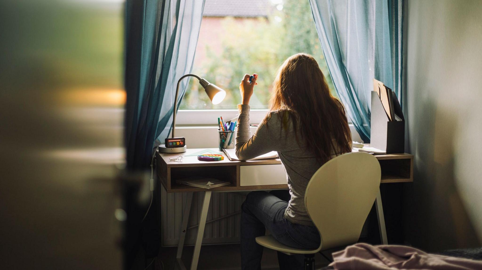 A teenage girl doing homework while sitting at desk in home. She is sat on a white chair and the lamp on the desk is on. There is a window nearby.
