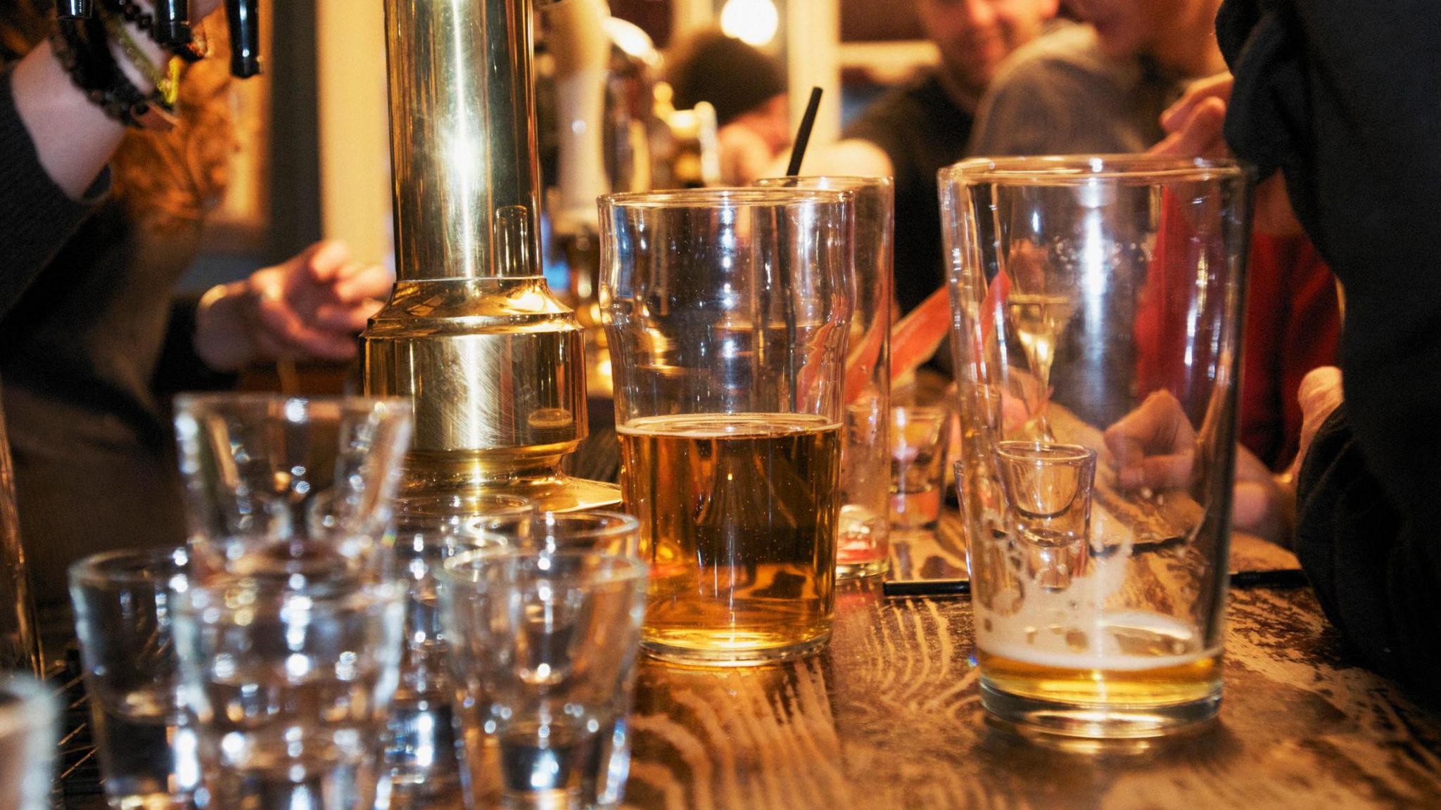 A collection of shot glasses and half-empty beer glasses on a bar with people in soft focus behind them.