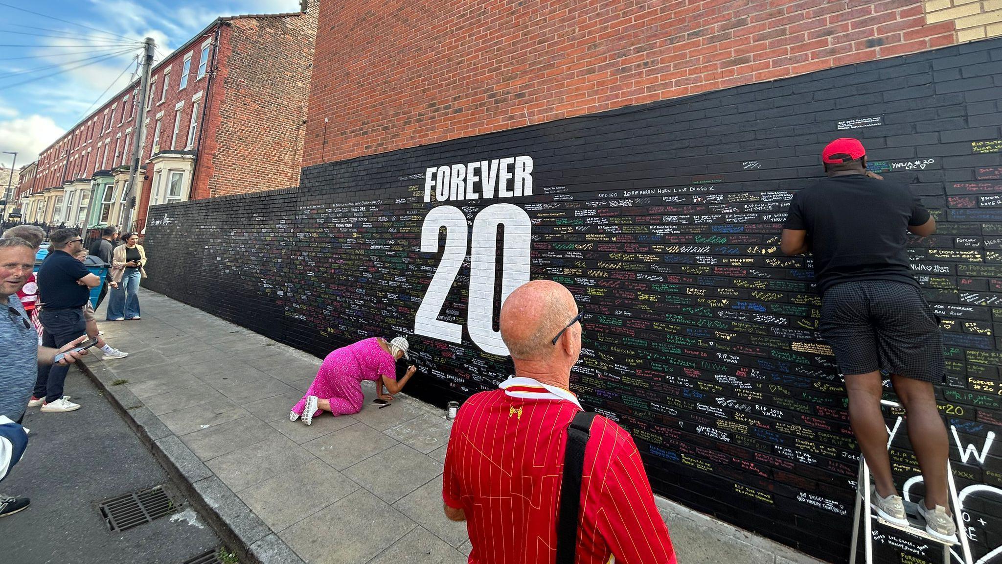 Fans stand in front of a mural on a gable end wall, consisting of the words 'Forever 20' in white letters against a black background. There are messages in different colours written on the brick, and a man on a stepladder and a woman crouching on the ground are pictured writing their own words.