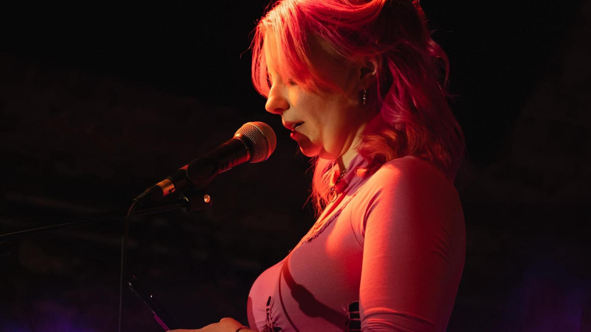 A woman is photographed from the side reading from a phone into a microphone. She has shoulder length hair and is lit up in a pink light.