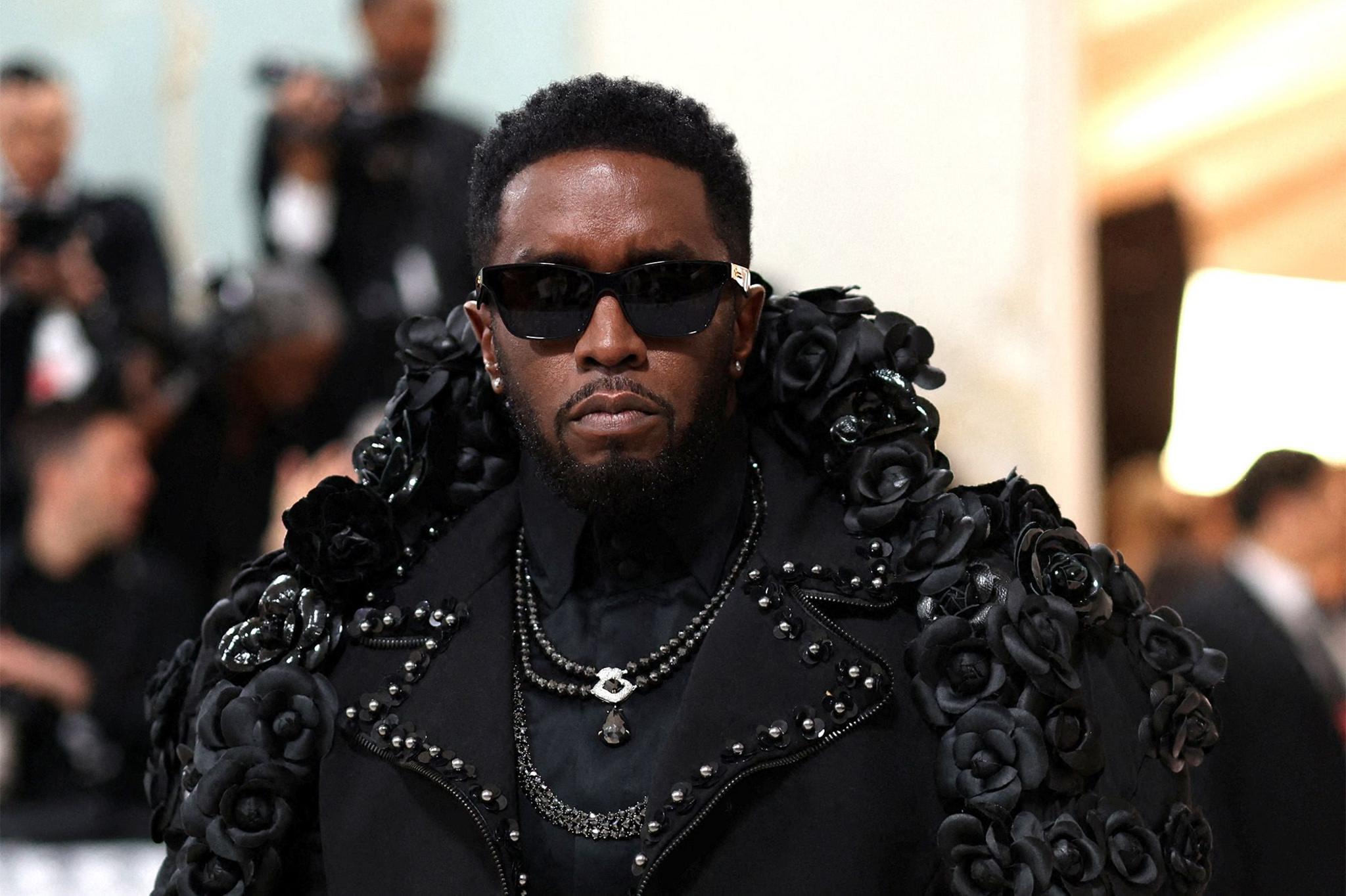 Sean Combs in an elaborate black outfit decorated with black roses and embellishments, surrounded by people in the background, some taking photos.