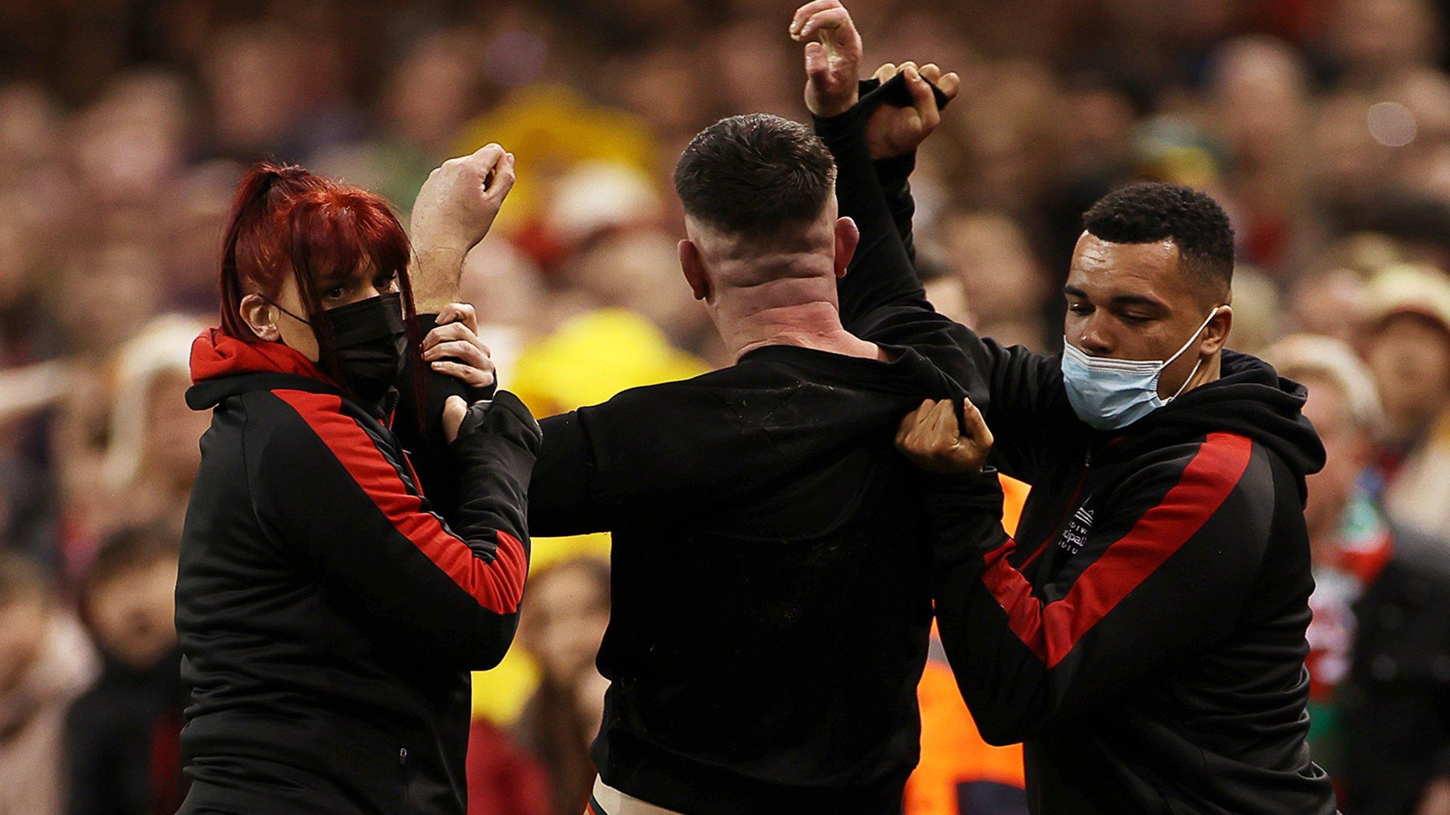Pitch invader is grabbed by the wrist. This image shows the faces of two masked security guards, but does not identify the pitch invader as his back is shown.