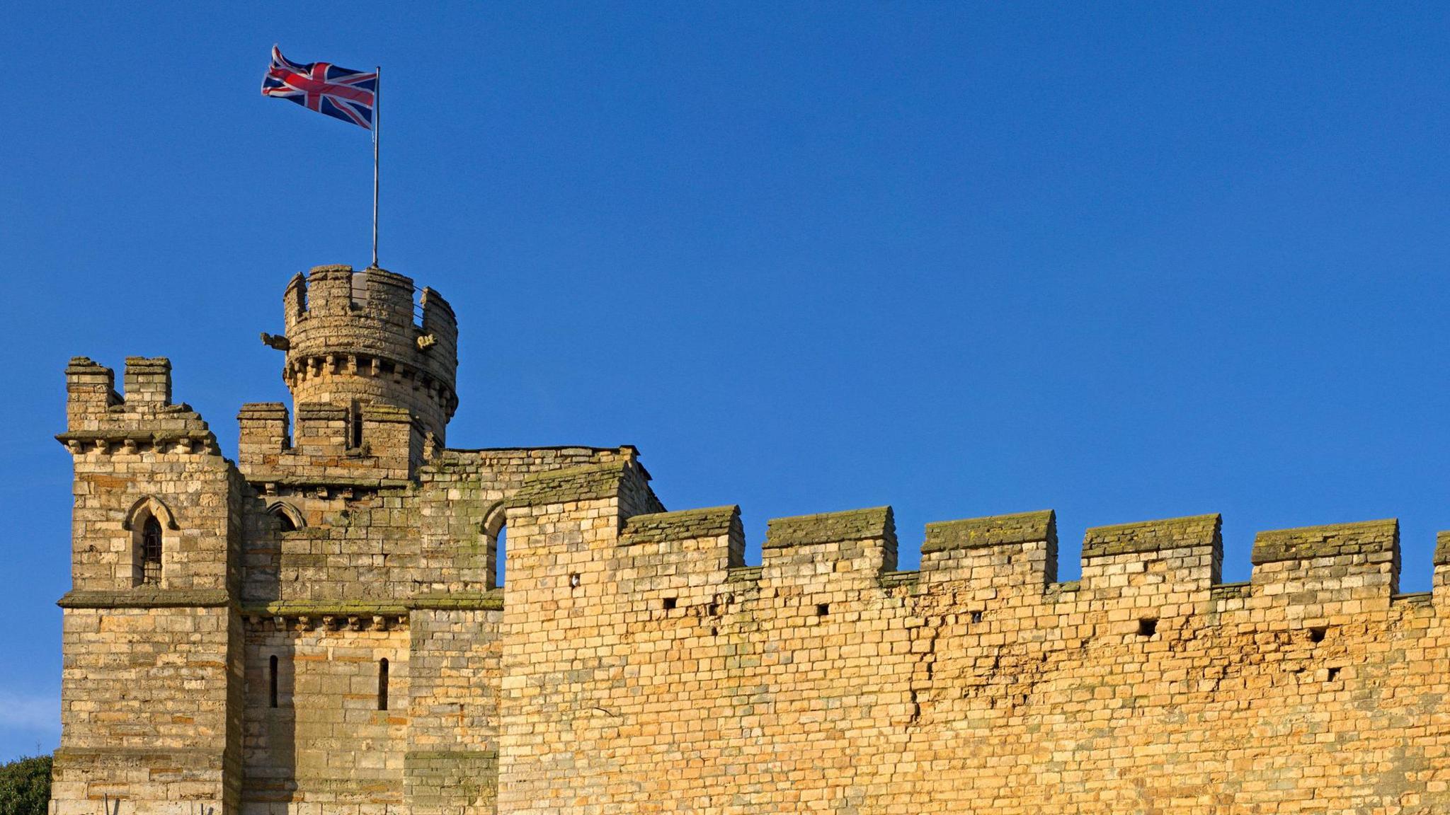 Lincoln Castle. The castle walls rise to a tower at the left of the picture from which a union flag flies. The sky is clear blue 