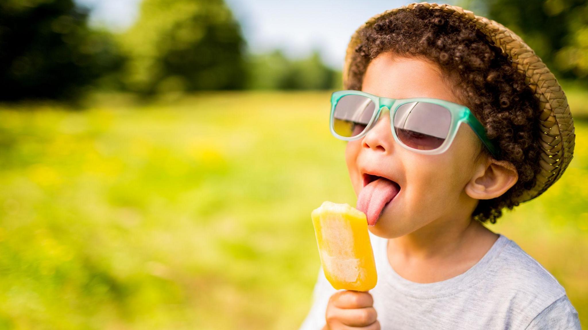 child in sunglasses and hat eating ice lolly