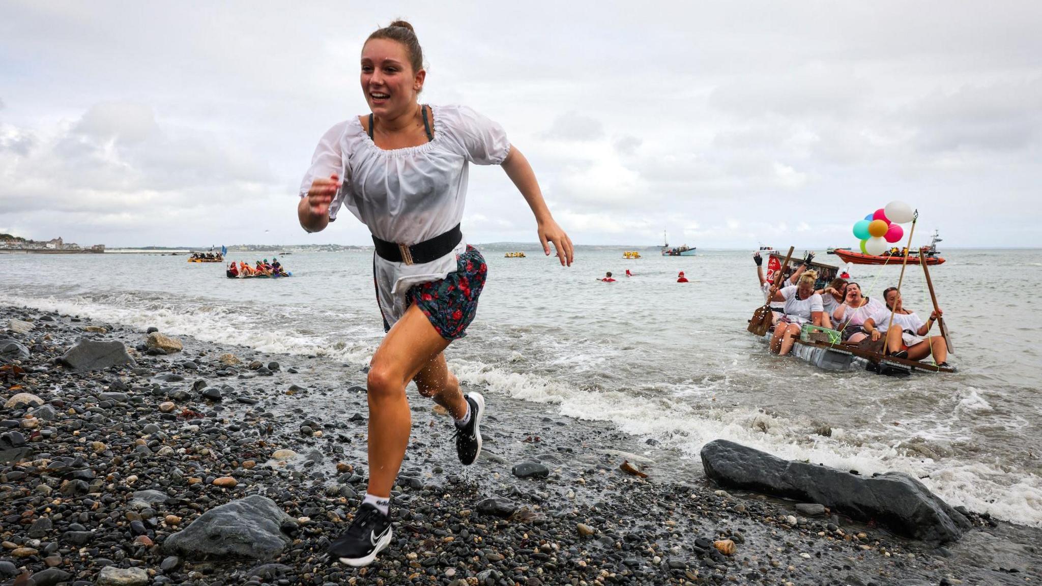 A women in wet clothing races up a pebbly beach with a raft of women in sisal costumes behind her in the sea.
