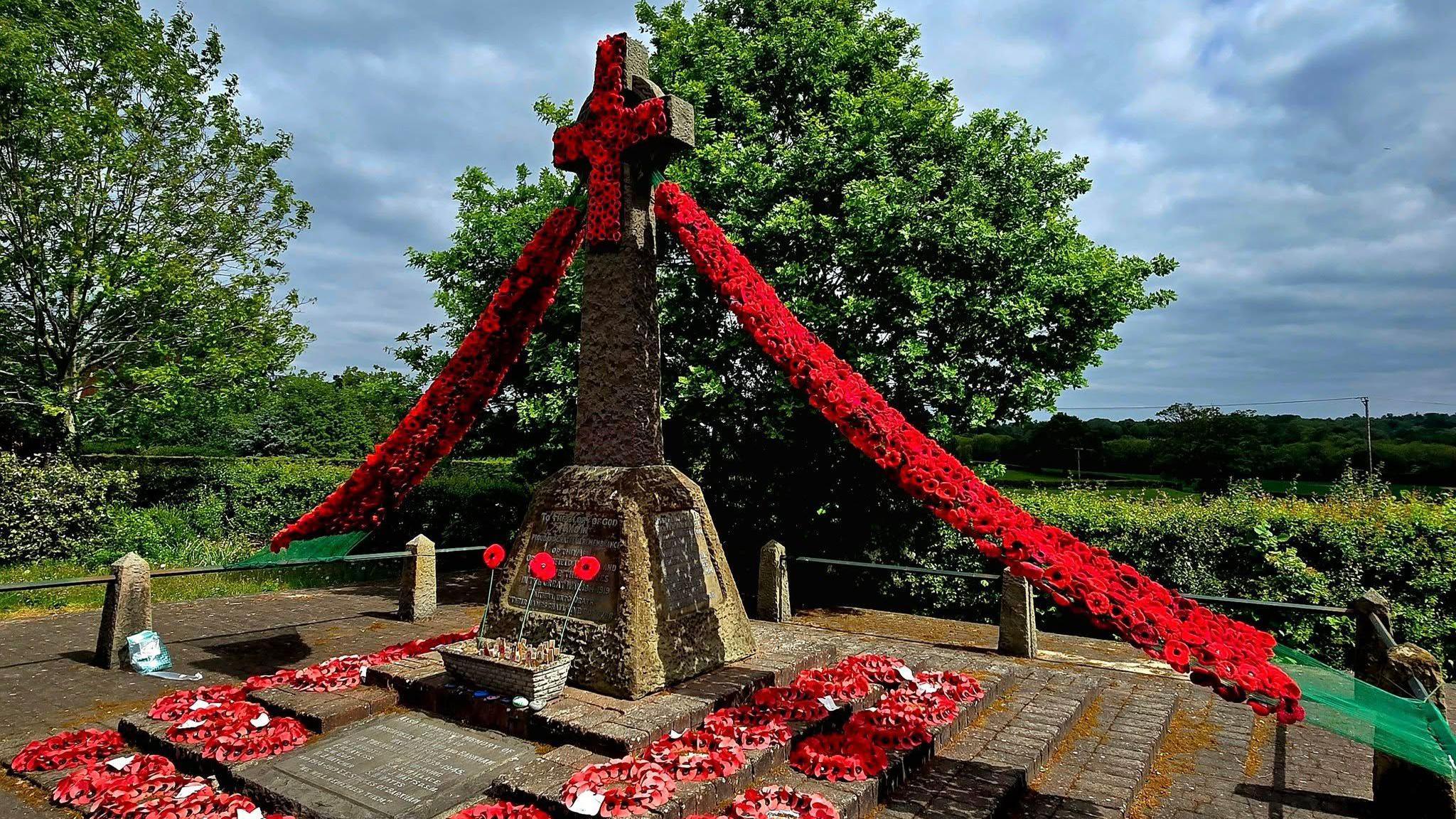 A stone war memorial surrounded by poppy wreaths and with a long garland of red poppies strung from its top down to two points on the ground either side.