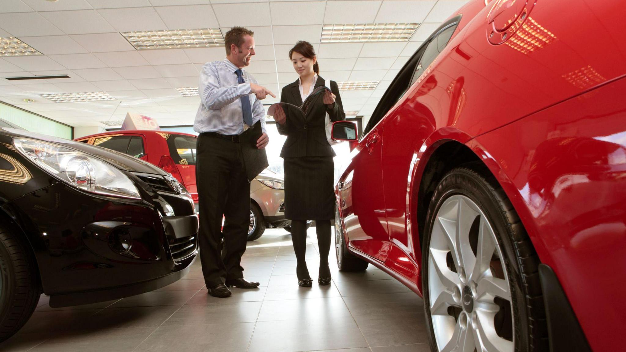 Man and a woman in smart clothing talk in a car dealership, standing between new cars