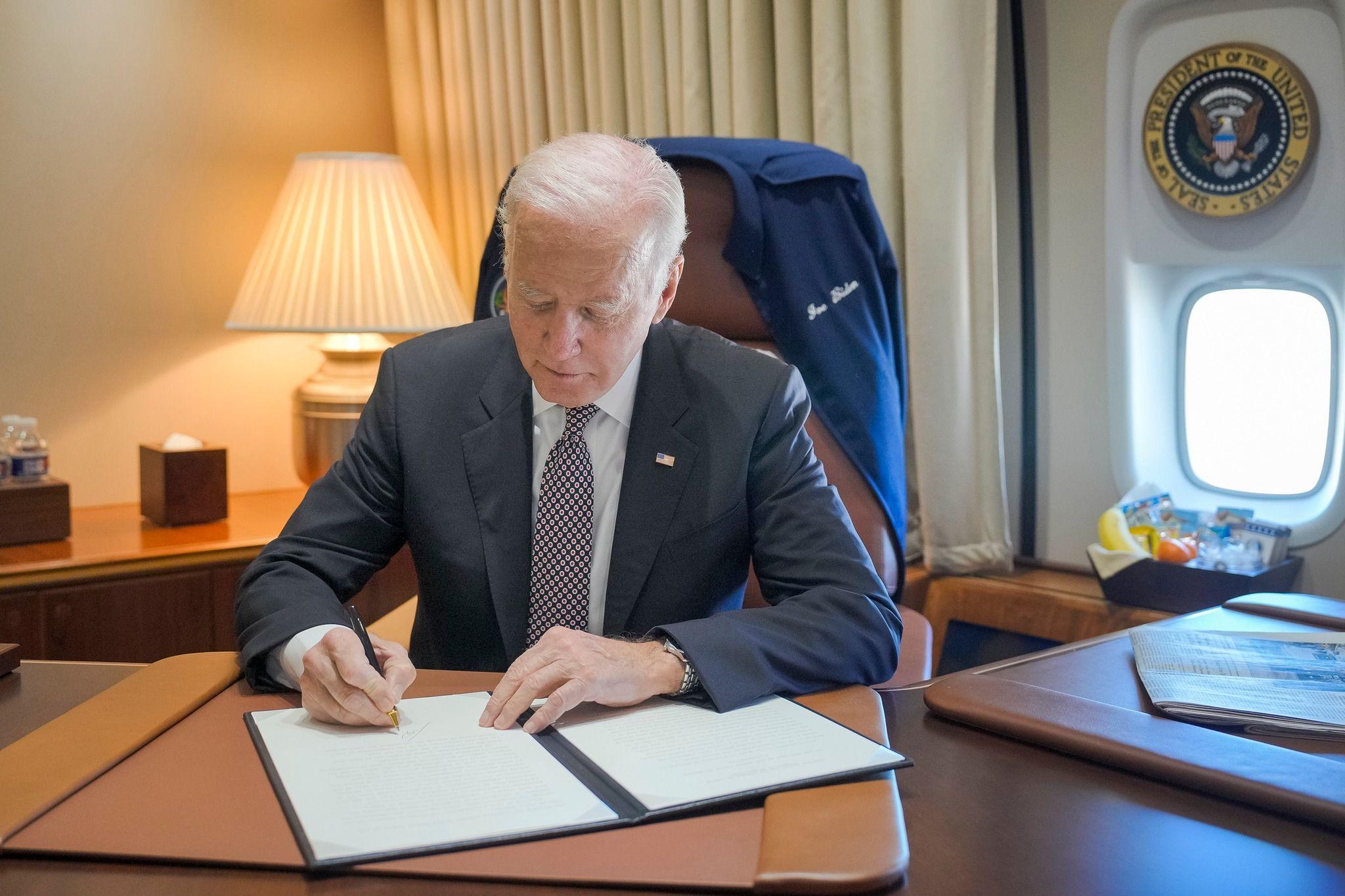 President Joe Biden sitting at his desk on Air Force One signing a pardon. He is wearing a US flag badge on his lapel. There is a blue jacket over the back of his chair with his name written on the front. The Seal of the President of the United States is above the plane window behind him. 