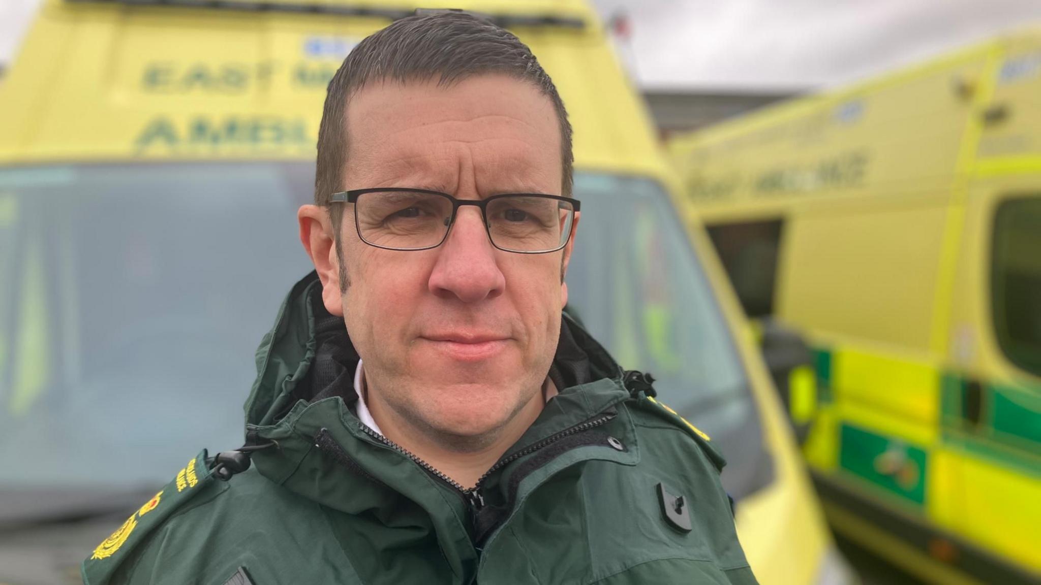 Director of Operations for EMAS Ben Holdaway dressed in a green jacket with badges on his shoulder. He is standing in front of two emergency ambulances and has black, metal-framed glasses on.