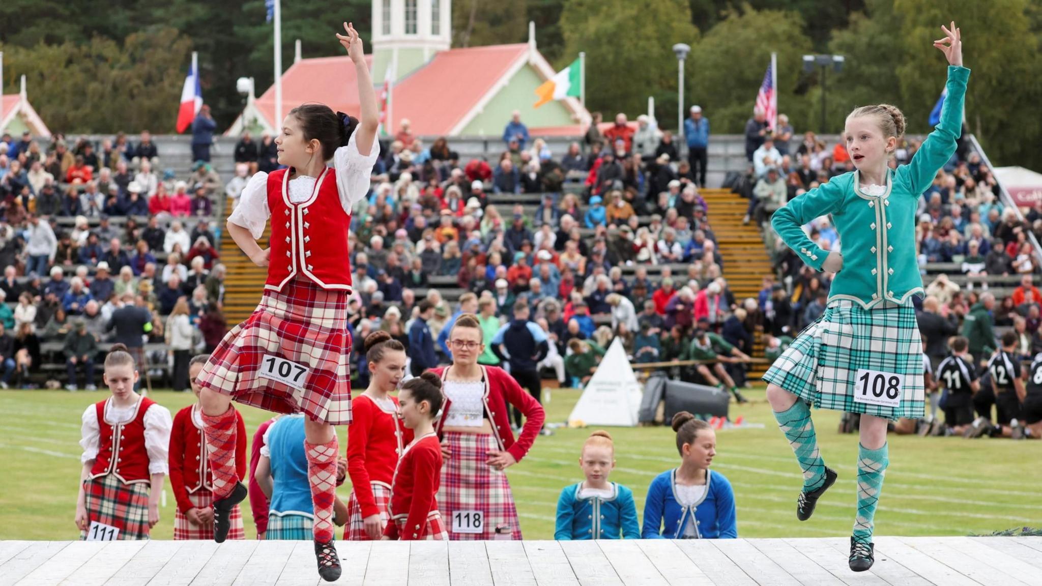 Two young girls taking part in a Highland dance display on a stage. They are dressed in traditional Highland dance outfits, including kilts and vests - one red and white and the other green and white.
There are a number of dancers standing behind them and spectators (blurred) further back.