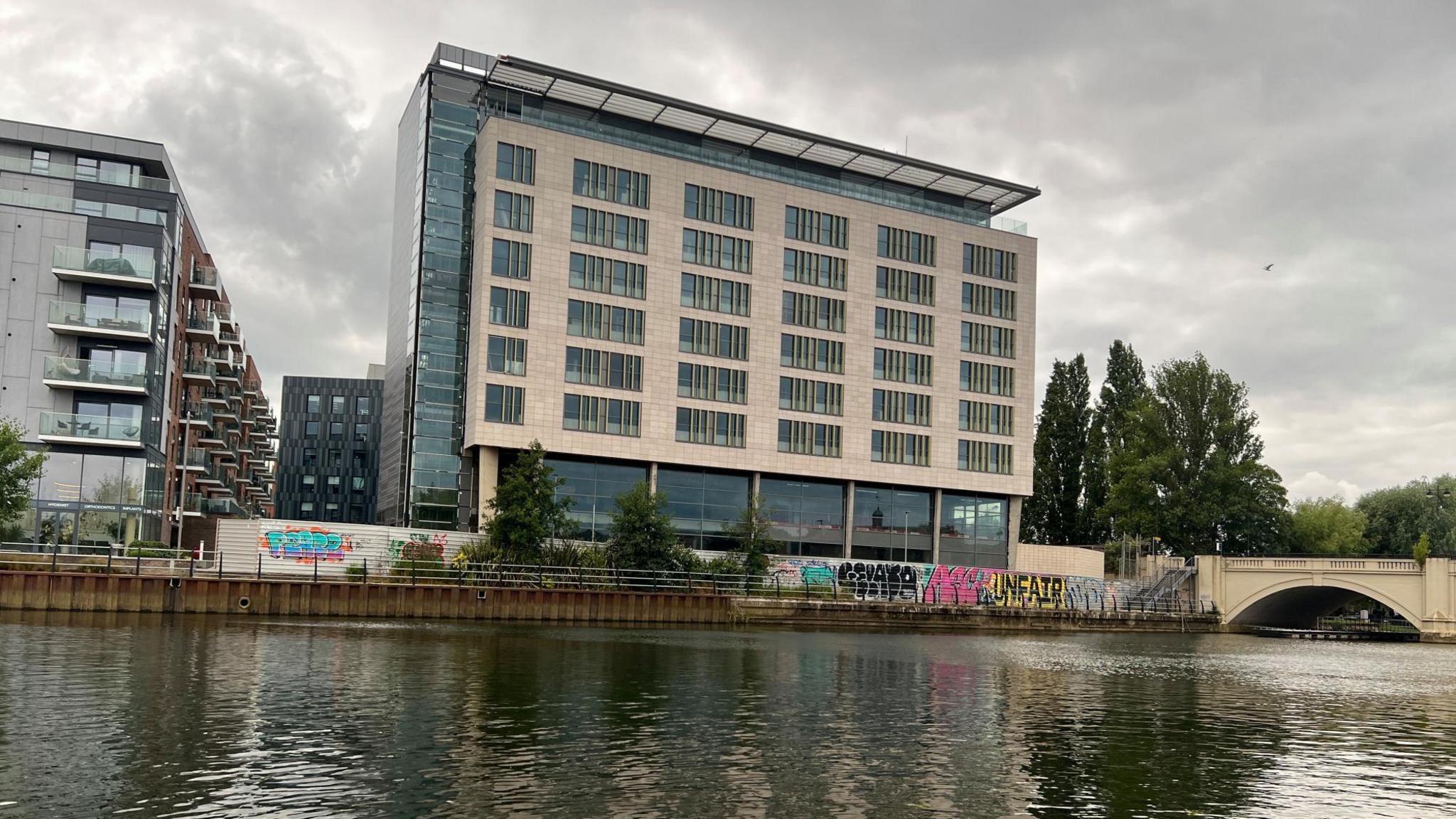 A six-storey building with beige cladding and glass sides is pictured from across a river