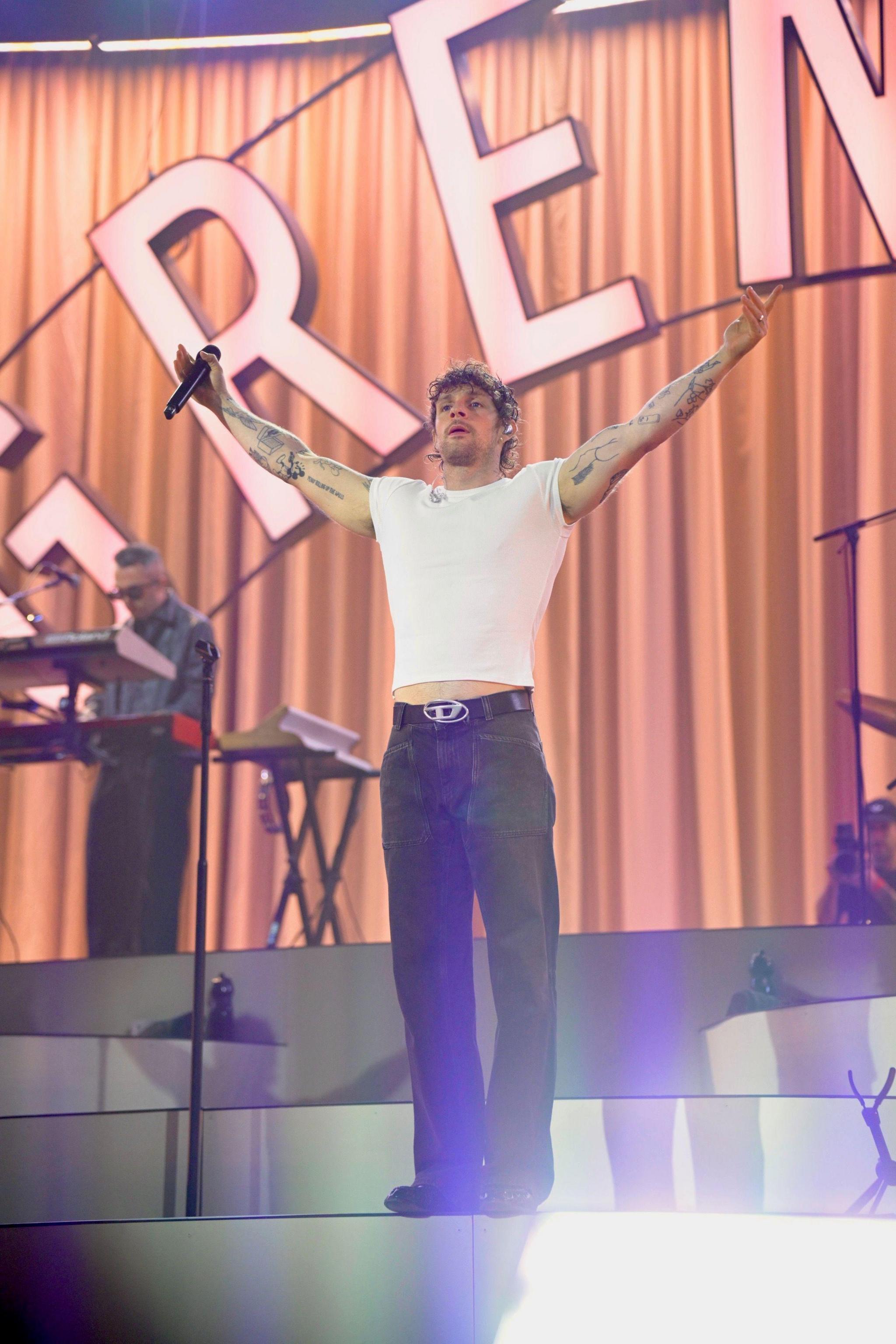 Singer Tom Grennan is wearing a white T-shirt and black trousers with his arms outstretched on stage with the word "Greenan" in large letters behind him. 