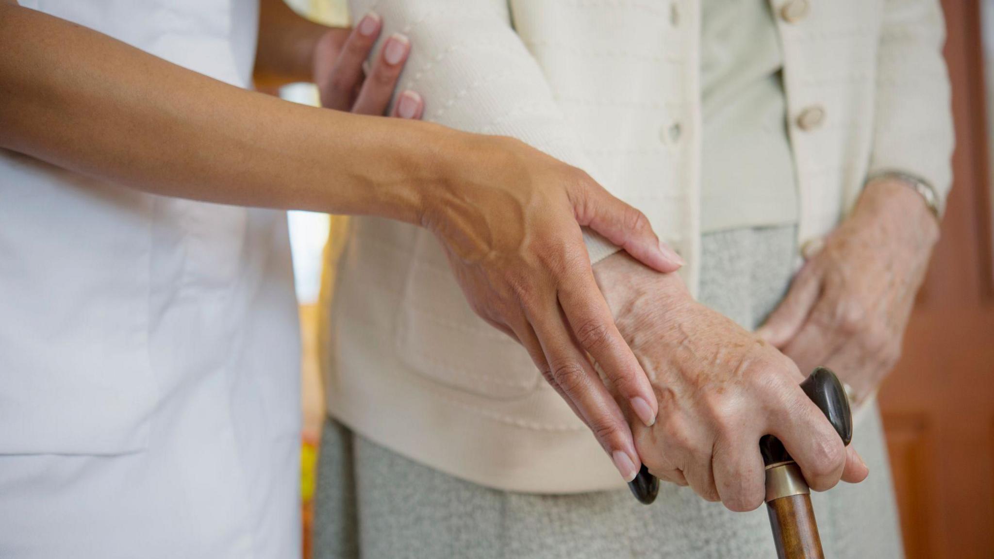 A close-up of an elderly lady's hand holding onto the top of a walking stick, with the hand of a carer helping her.