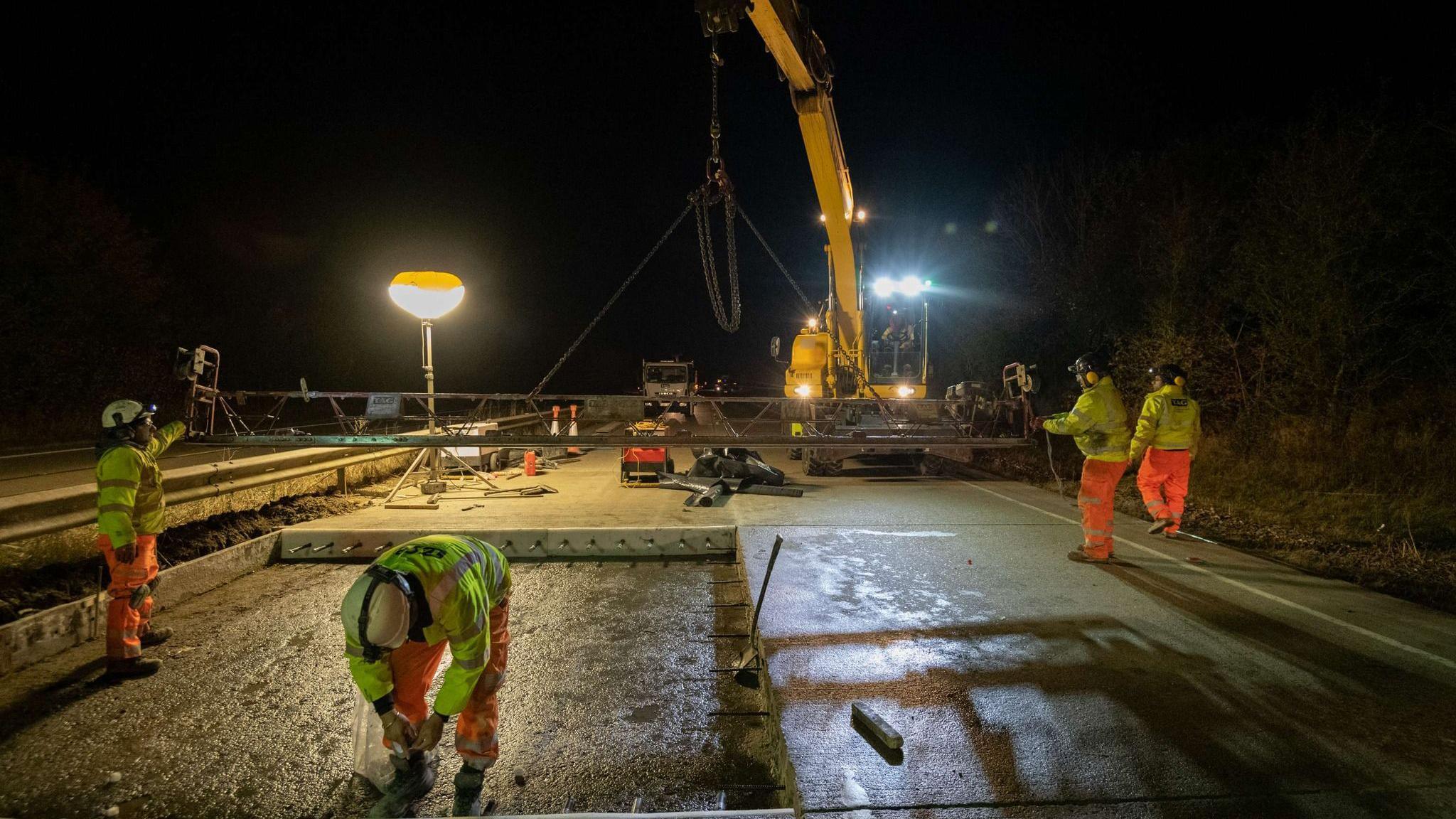 Workers wearing high-vis and helmets are stood on a road where a large section of the concrete has been removed.