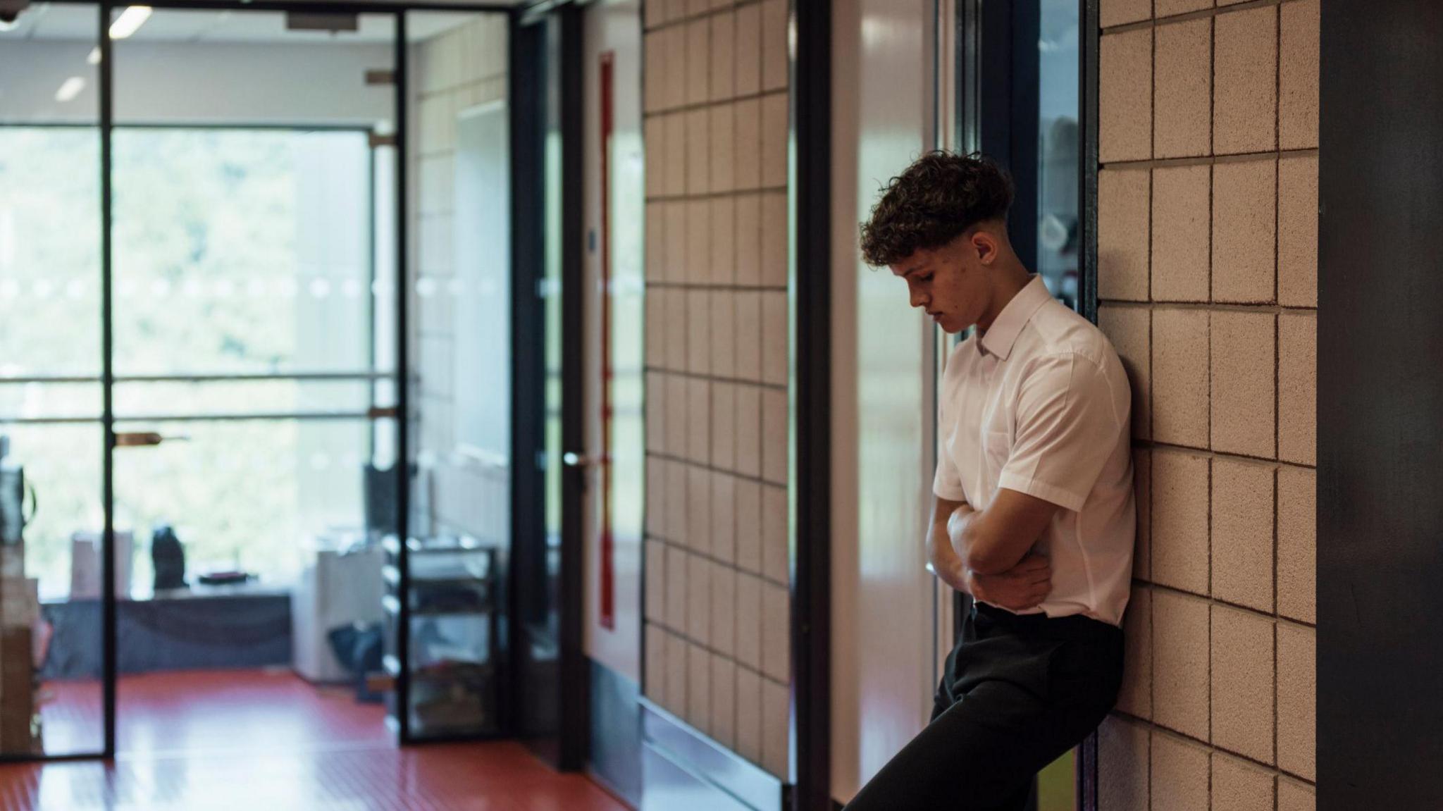 A school boy looks forlorn stood with arms folded outside a school classroom