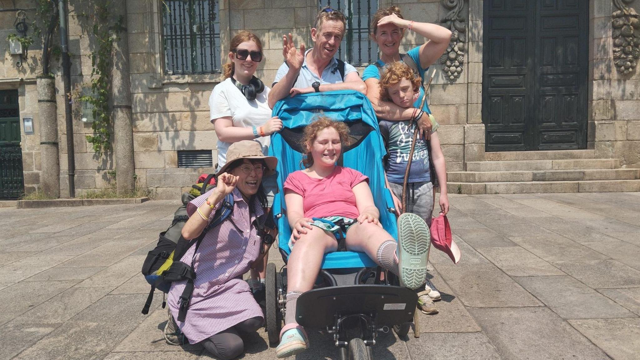 A family in front of an old building. In the middle is a teenage girl in a buggy, surrounded by her mum and dad and a brother and sister.