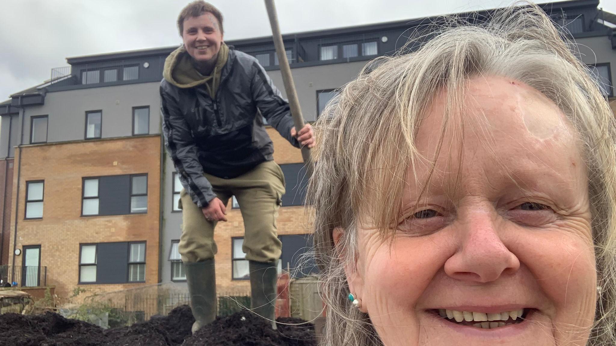Sara, front right, taking a selfie photo, while Noah, rear, stands and smiles. He is holding a spade, and the pair are standing in front of an apartment block. Sarah is older and has grey hair, Noah has a black jacket, green trousers and wellies on