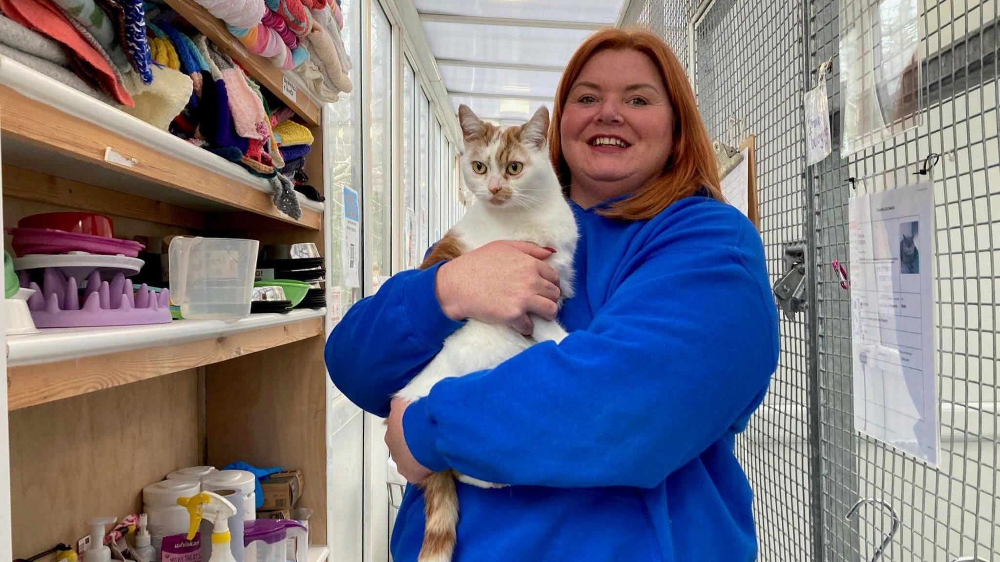 A ginger and white cat with a pink nose being held by a woman with reddish/brown hair wearing a blue fleece.