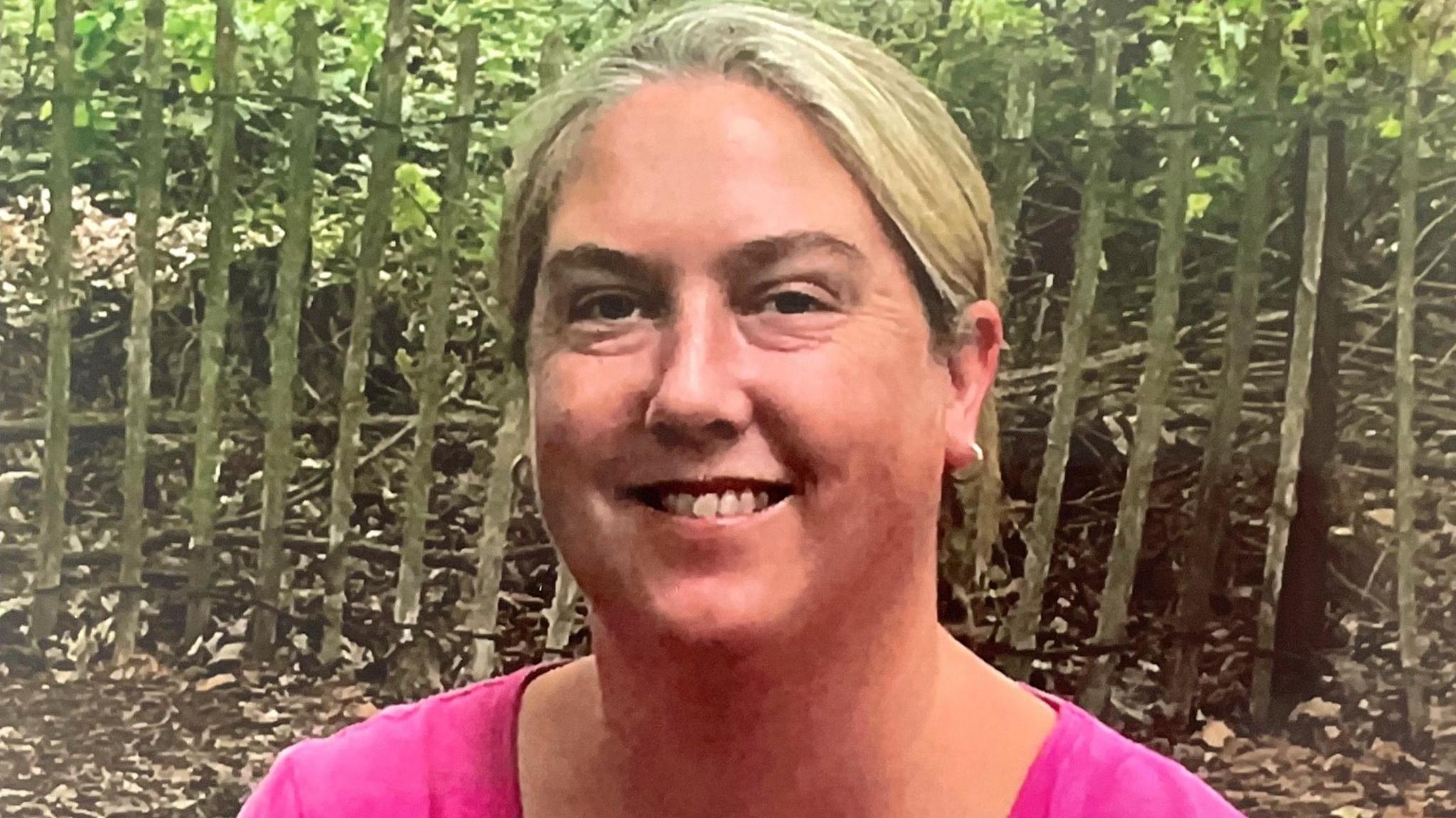 Julie Hurn has blonde hair tied back and is wearing gold hooped earrings and a fuchsia pink top. She is smiling at the camera and photographed outdoors. 