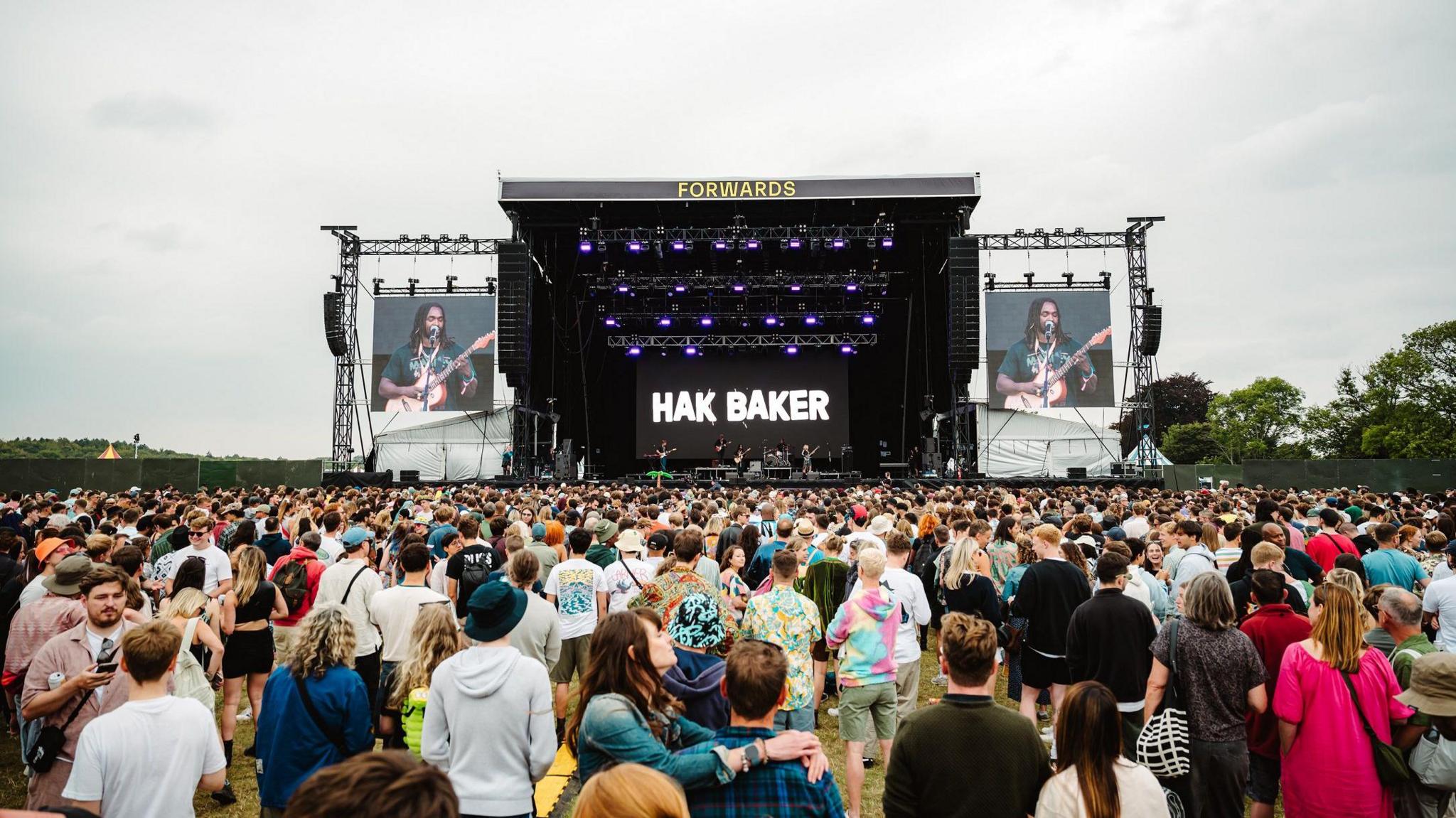 A large crowd is gathered in front of the main stage at Forwards Festival in Bristol. On the big screens on either side of the stage a main is seen playing a guitar and singing.