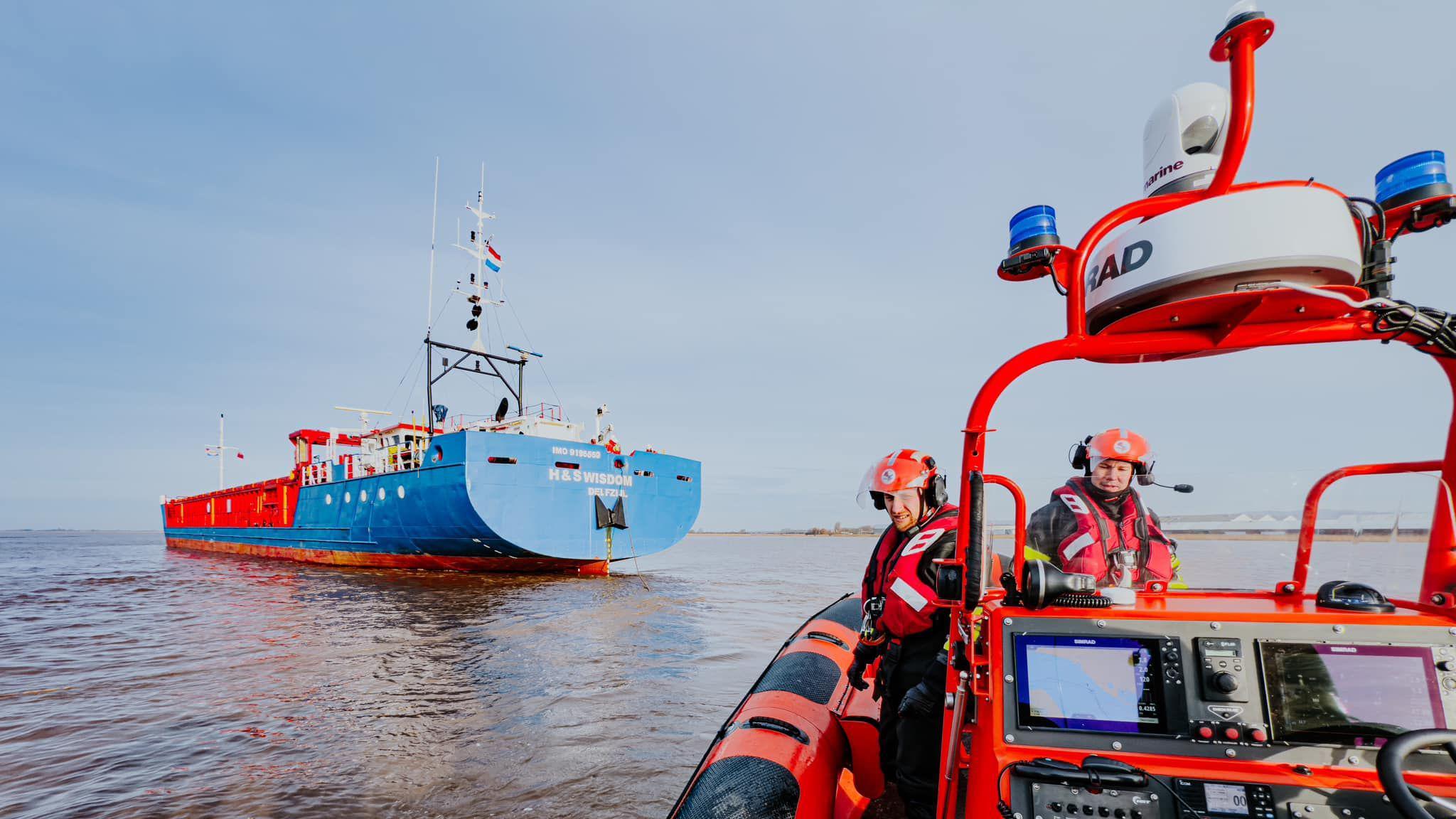 A Humber Rescue lifeboat can be seen on the right hand side out at sea with two men operating it and wearing red lifejackets and helmets. A large vessel can be seen in the distance on the left.