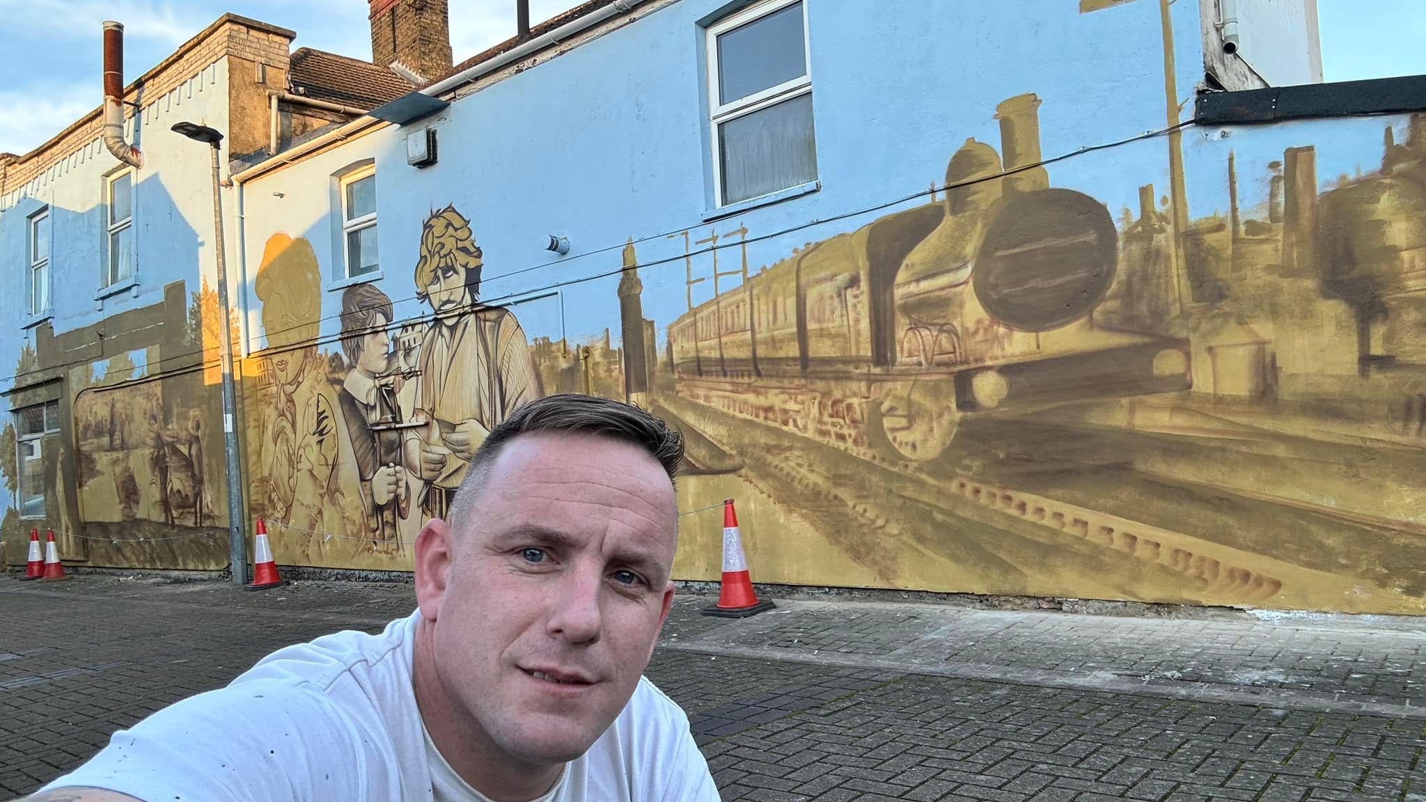 Nathan's selfie in a white T-shirt in front of his mural wall