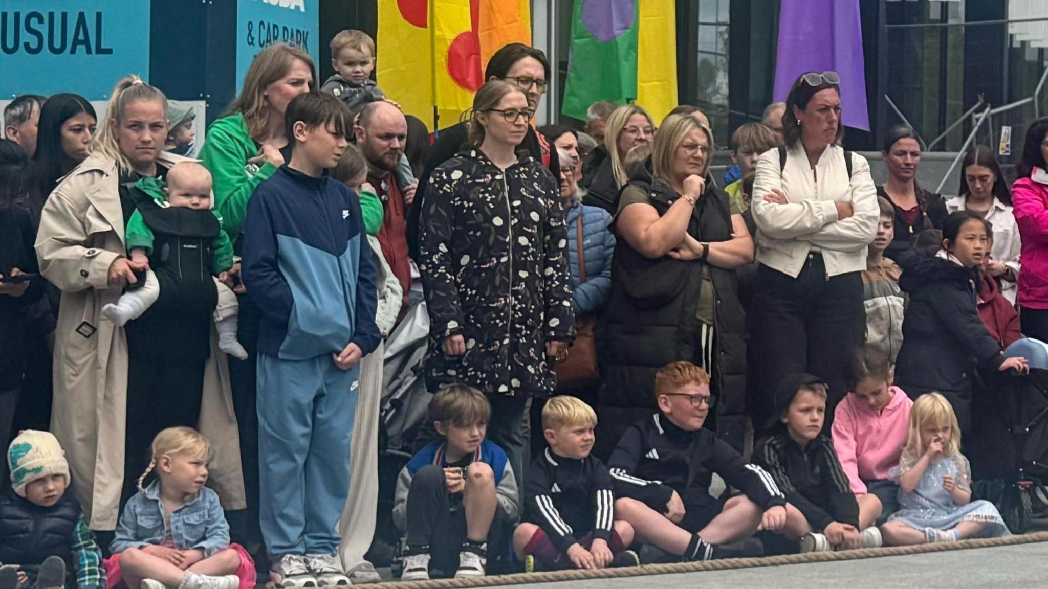 About 10 young children sit on the ground watching performances with adults standing up behind.