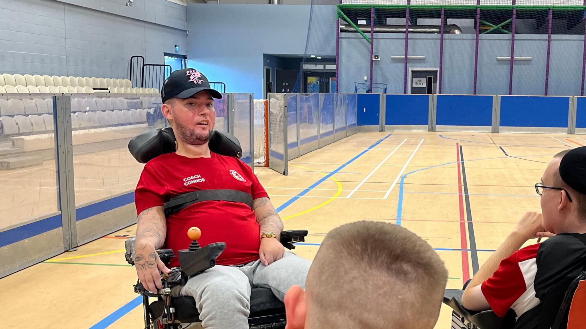 Connor Rumney is sitting in a wheelchair on a sports court. He is speaking with others and is wearing a black cap and a red sports t-shirt.