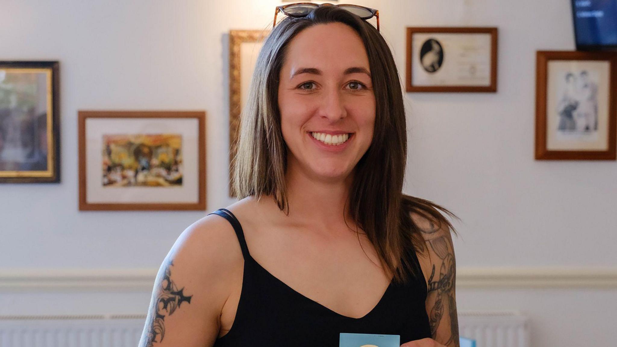 Amie Gray stands in a room with pictures on the wall. She has long, straight dark brown hair. Her black vest top shows tattoos running down both arms.