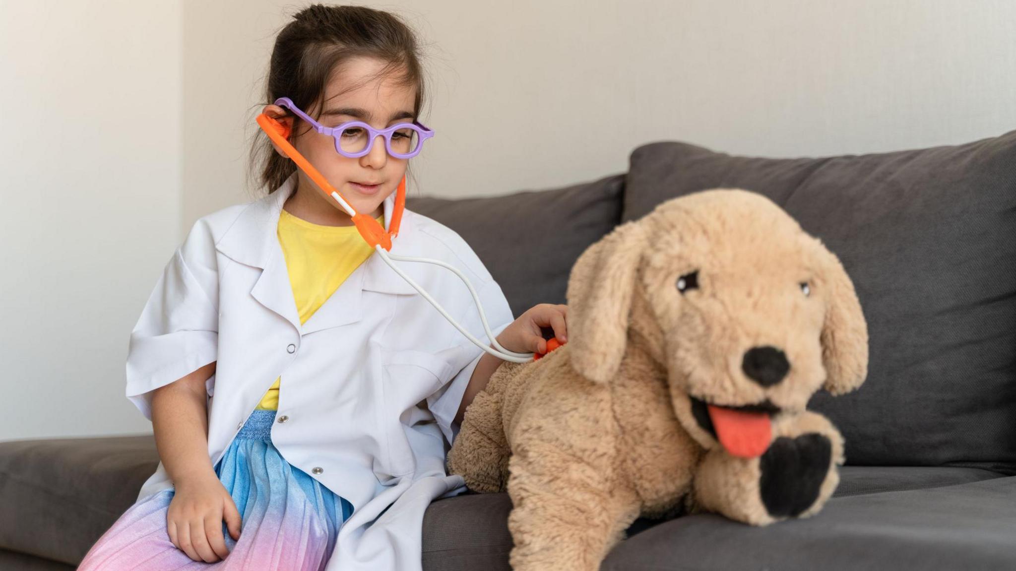 a girl wearing a dressed up as a vet in lab coat a stethoscope and glasses sits on a sofa and pretends to listen to her stuffed toy dog's heartbeat