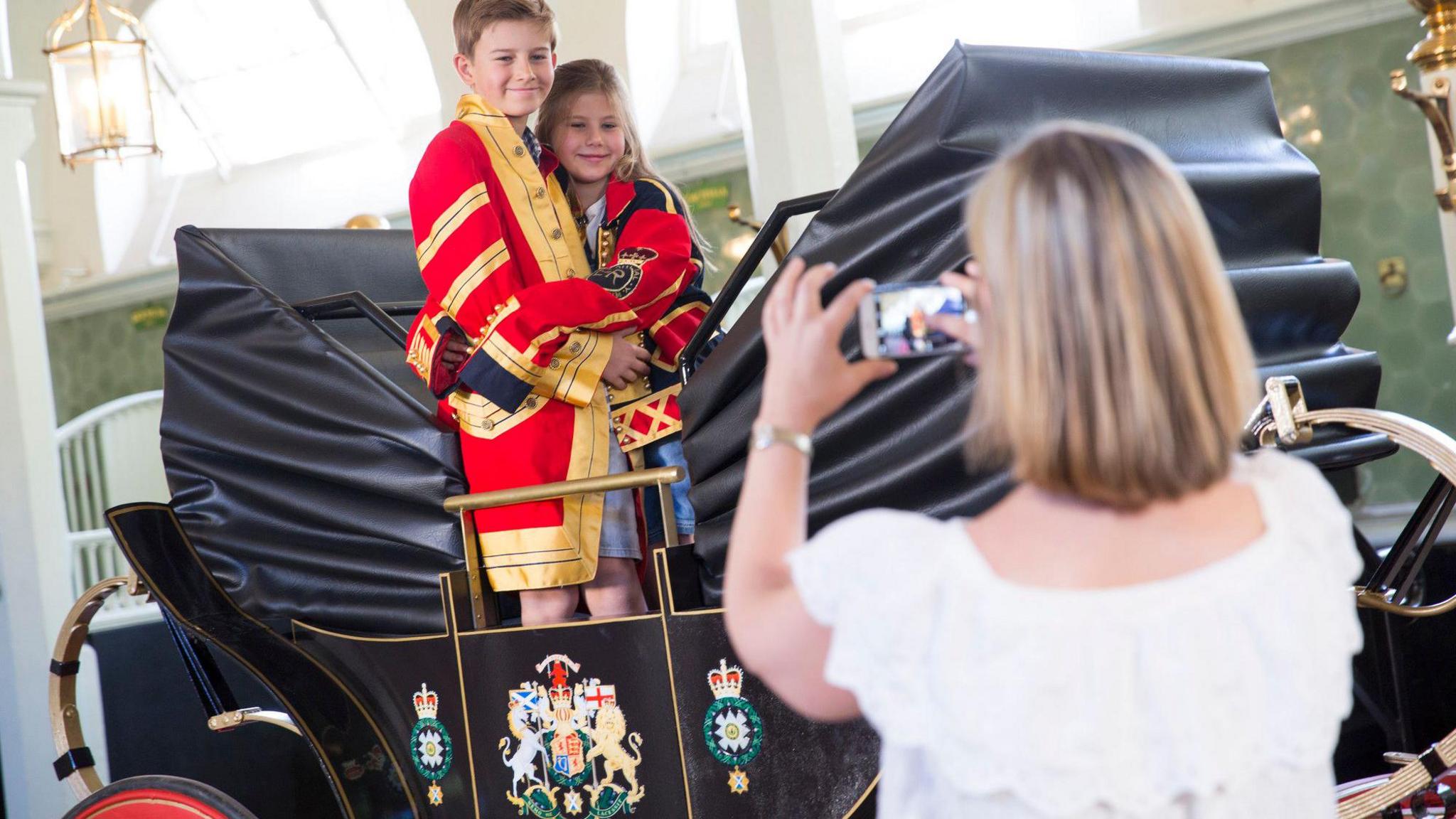 Two children wearing a livery jacket stand in a coach as a woman takes a photo of them. 