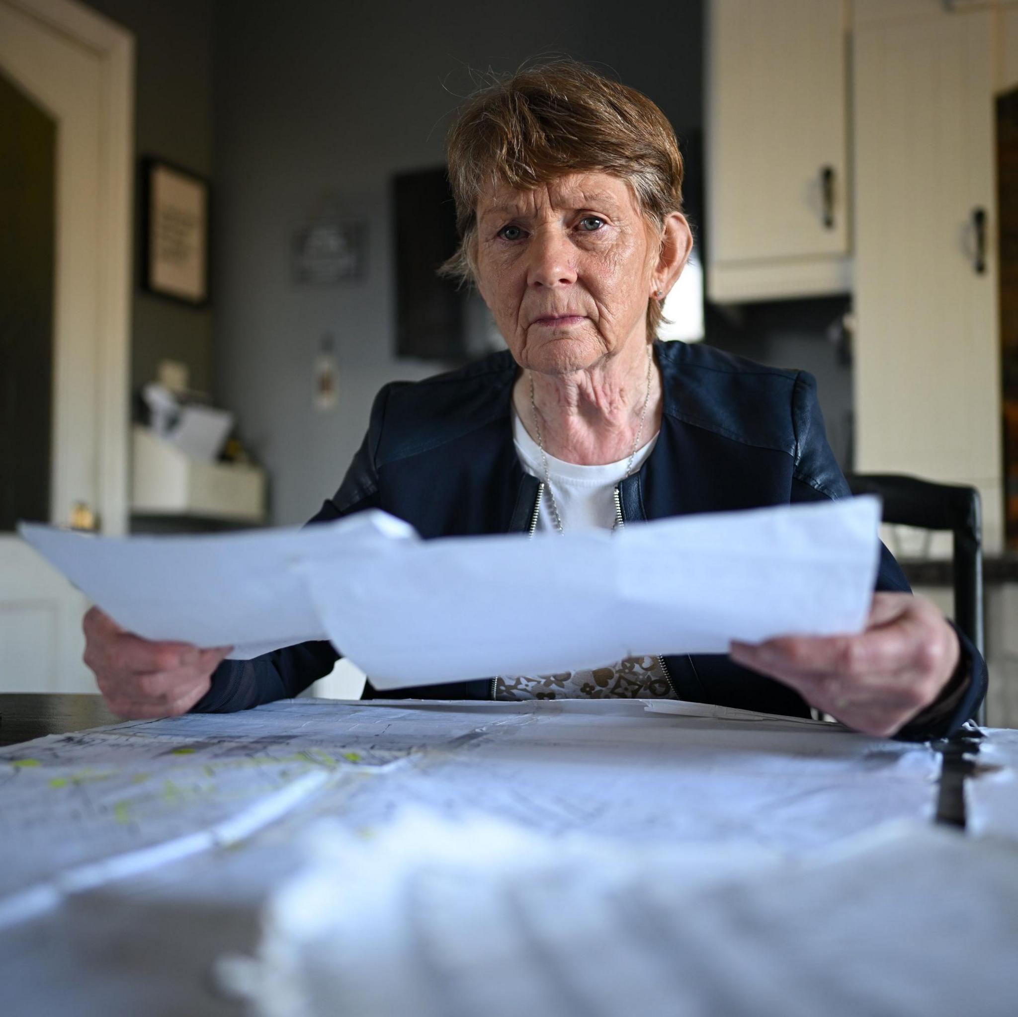 An older woman sits at a kitchen table. She is holding a sheet of paper in each hand. On the table in front of her are more documents and sheets of paper. She has short brown, greying hair and is wearing a white top and a blue jacket. In the background there are kitchen cupboards and a kitchen door which has two glazed windows set into it.