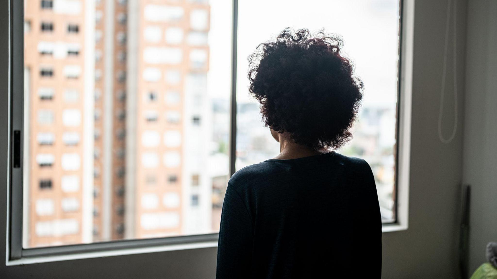 The silhouette of an unidentifiable woman with her back to the camera, staring out of a window.