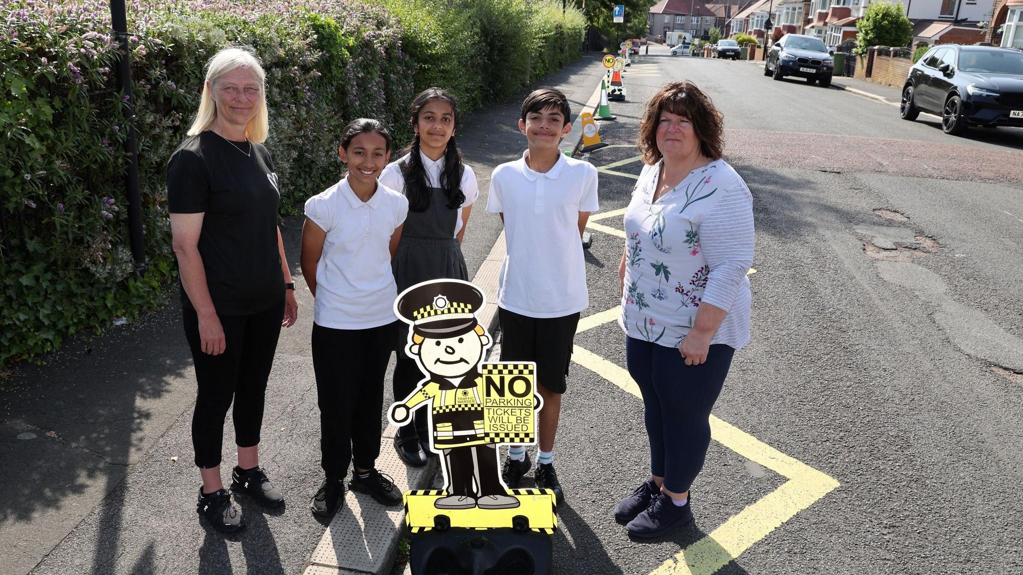Councillor Joanne Laverick with two girls and a boy, and Claire McKinney. They are standing in a no-parking zone alongside one of the "parking buddies" - a cartoon-like image of a policewoman holding a sign which reads "No parking, tickets will be issued".