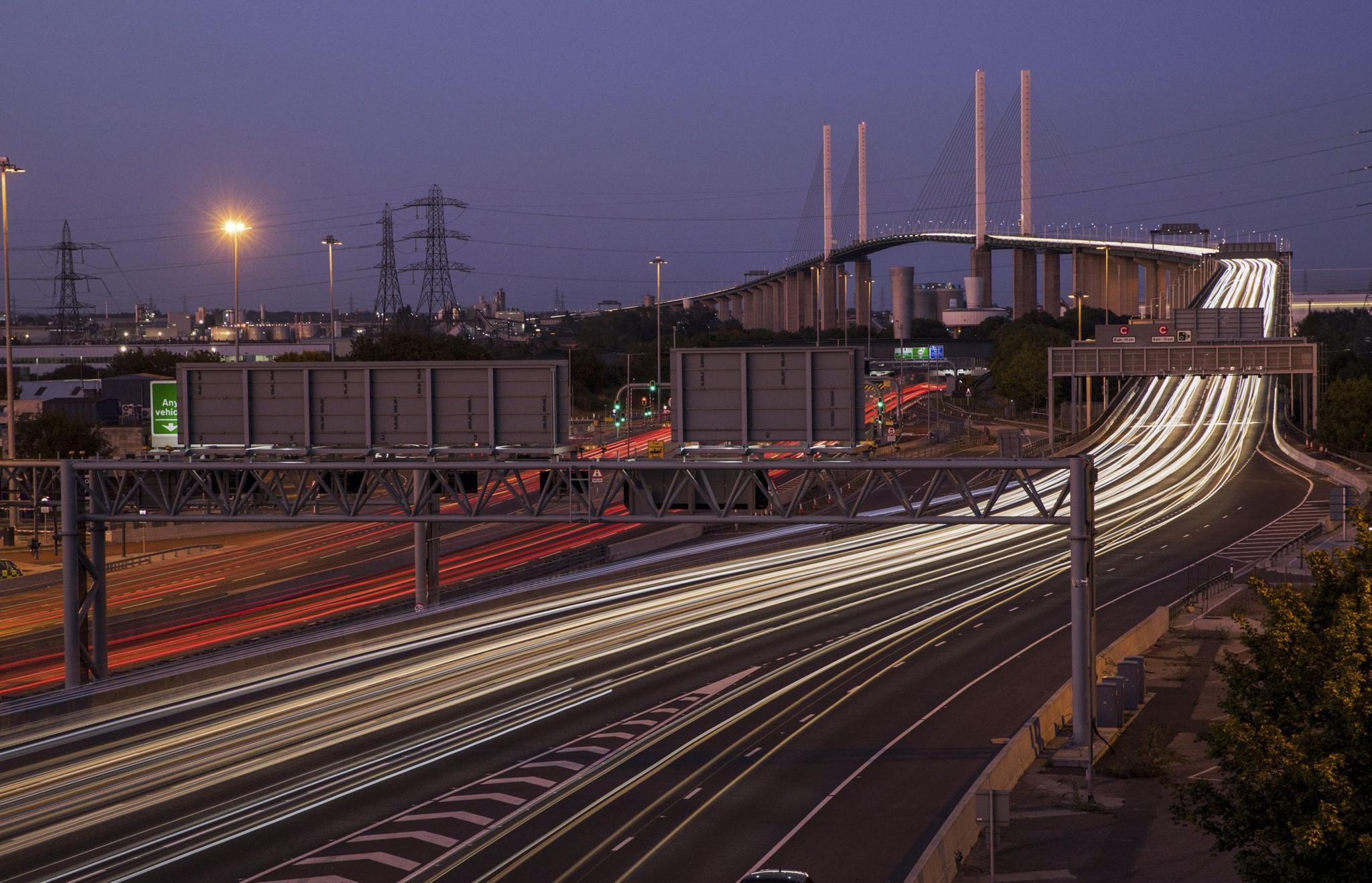 A view of the Queen Elizabeth II Bridge at night. There are two separate light trails from vehicles travelling over the bridge, one red from the back lights of vehicles, the other white, from their headlights.