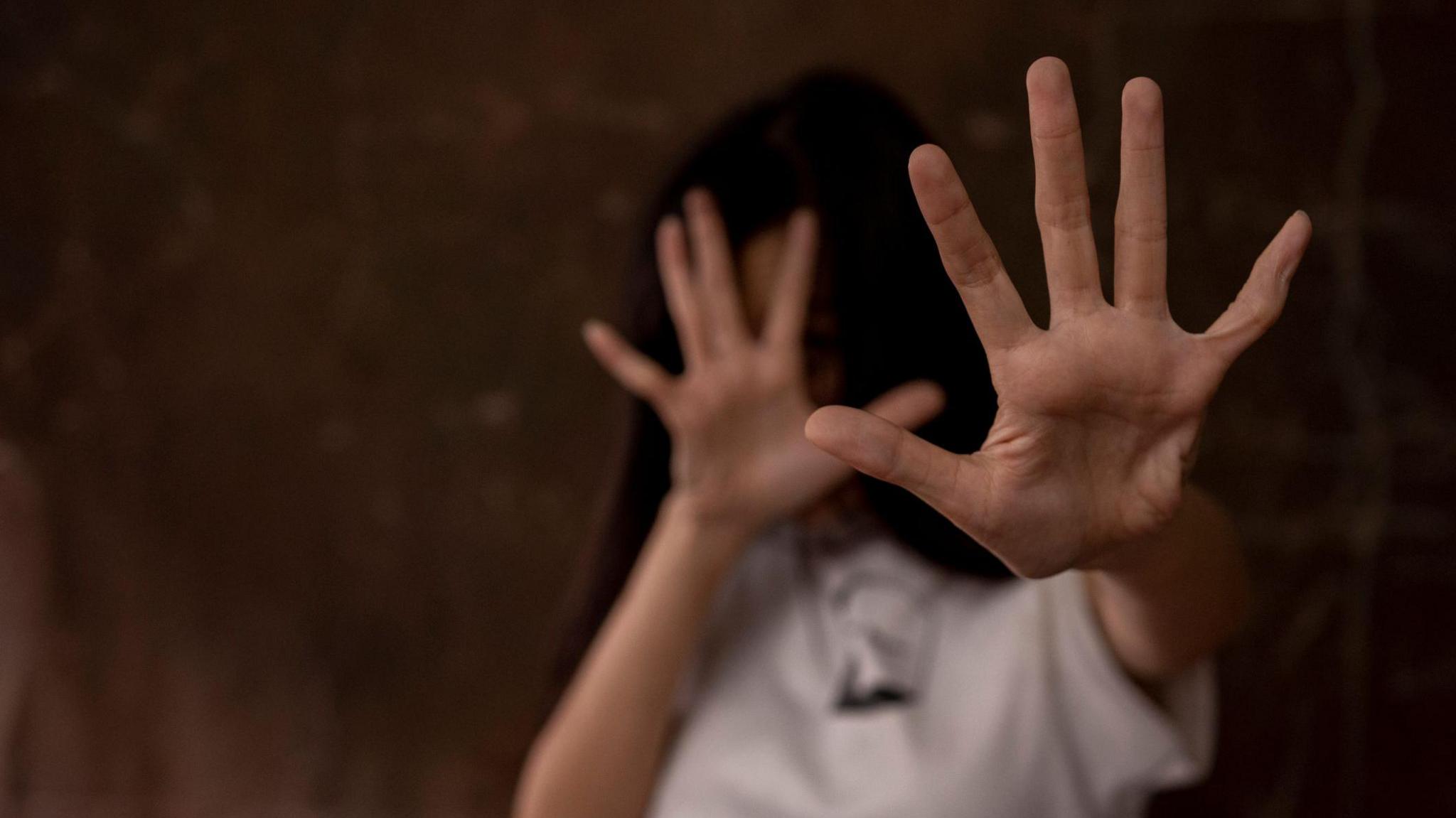 A young women holds her hands out in a defensive position. Her face if blurred out. she has dark hair and a white t-shirt