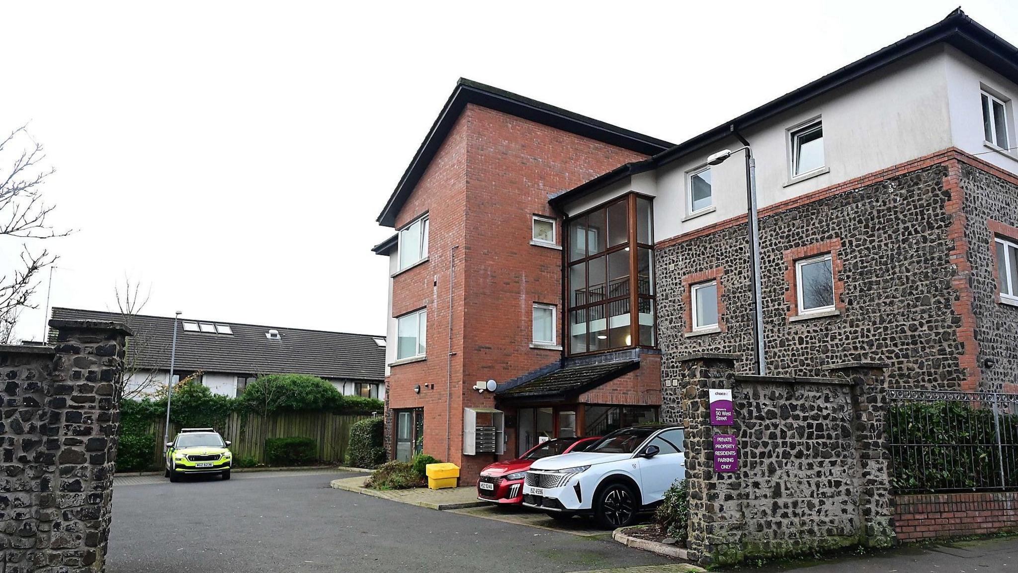 The flats on West Street in Newtownards seen in daylight.   The exterior of the three-storey building is a mix of red brick, cut stone and white render.  It has a angled roof on one side and a glazed atrium housing a central staircase.  A police car remains parked outside. 