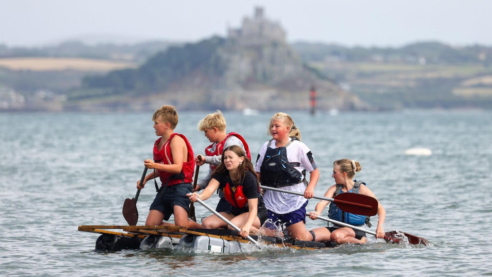 A team of young boys and first on a makeshift raft with St Micheal's Mount behind them. One young girl looks particularly anguished as she rows.